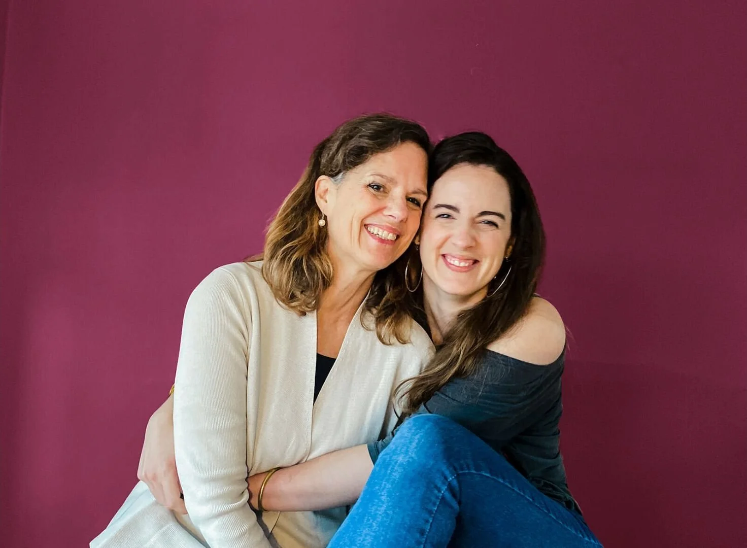 Two women smiling and embracing against a purple wall, one with shoulder-length brown hair wearing a cream-colored blazer, and the other with long dark hair wearing a black off-shoulder top and hoop earrings.