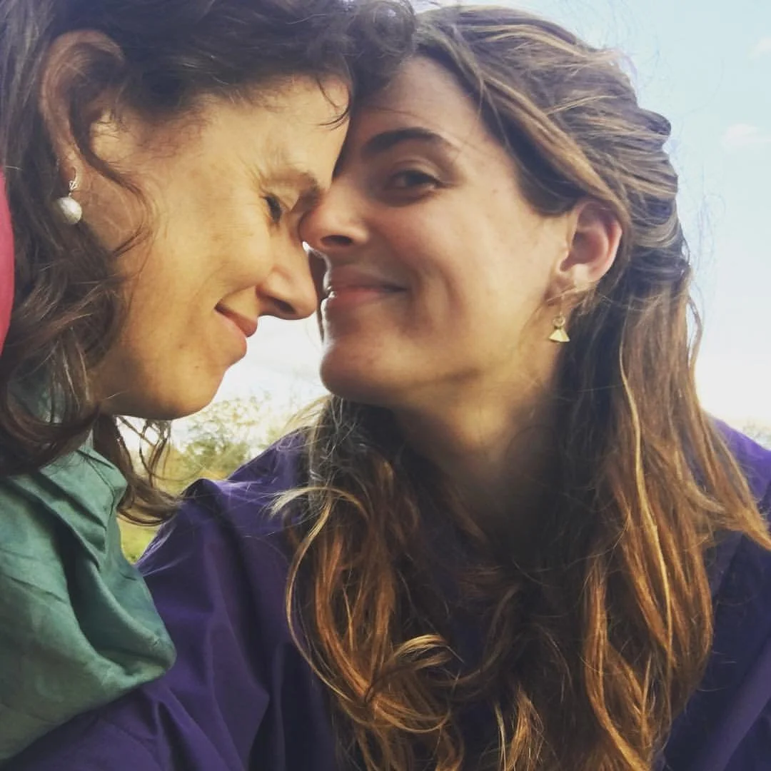 Two women with long, curly hair, close together with foreheads touching, outdoors under a blue sky, sharing a moment of affection.
