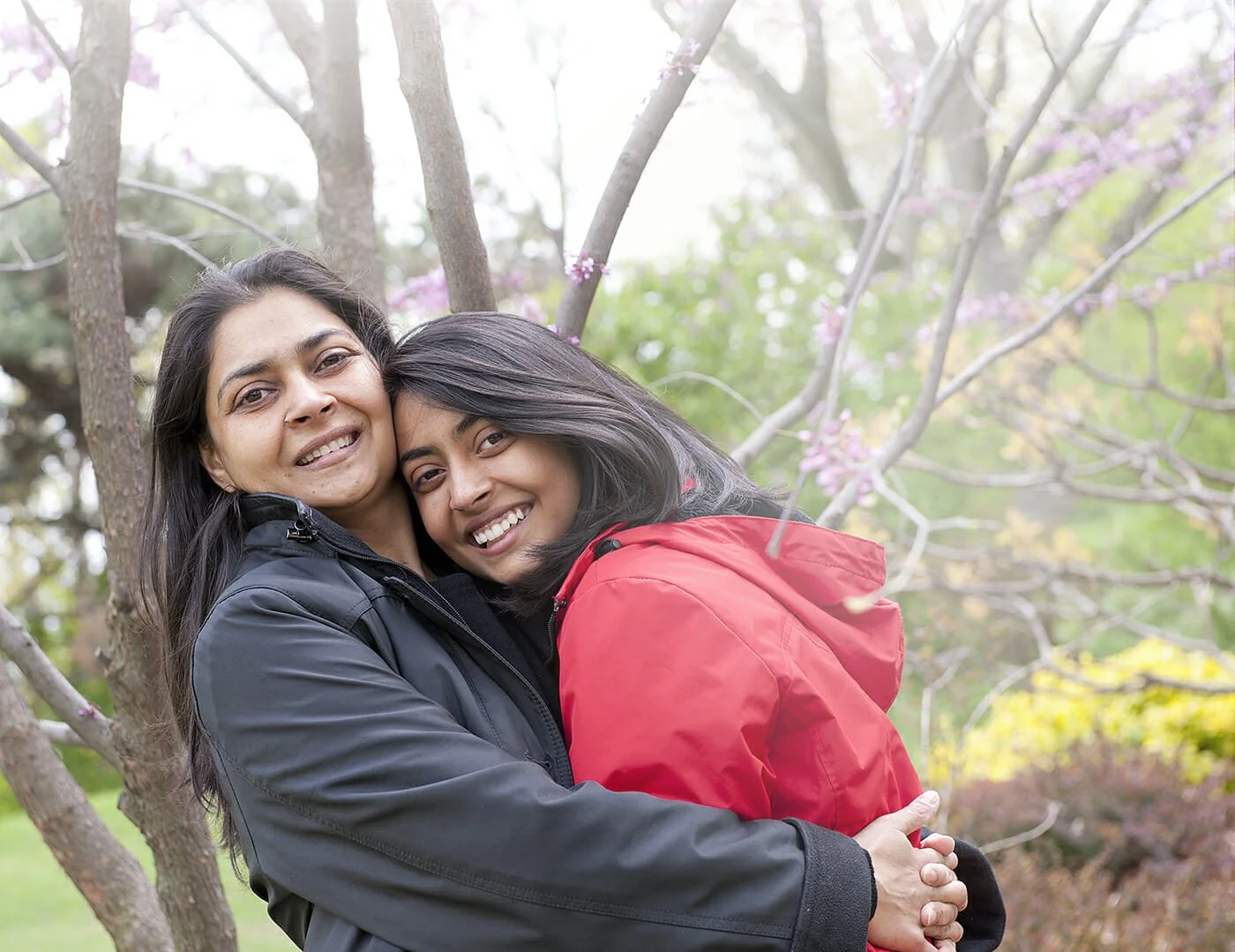 A woman and a child hugging outdoors in front of a tree with pink blossoms, smiling at the camera.