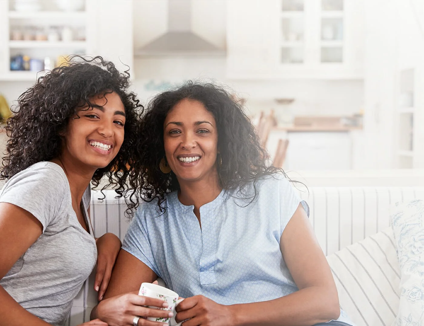 A teenage girl and a mother sitting on a white couch smiling at the camera, one holding a teacup.