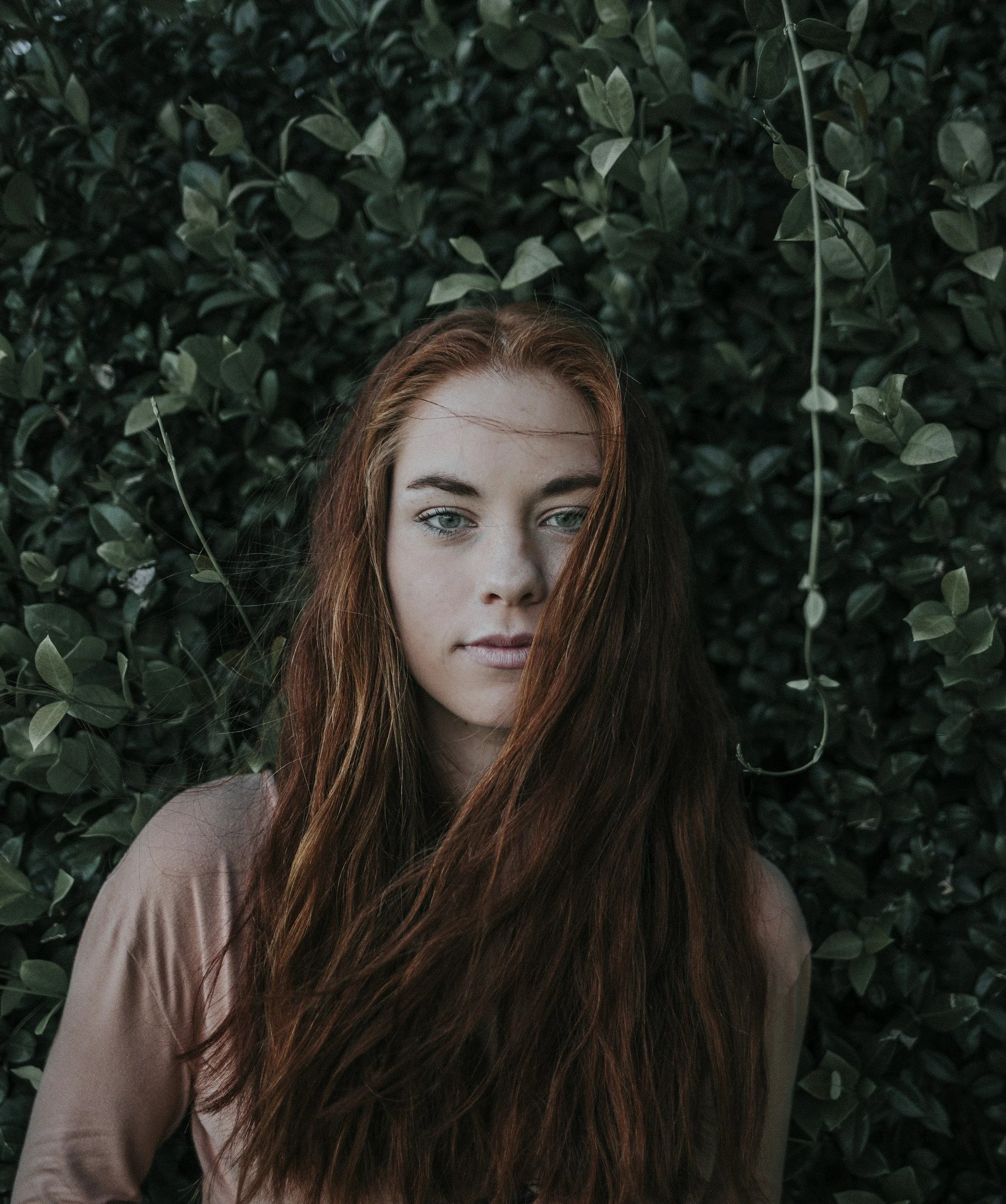 Portrait of a woman with red hair and blue eyes standing in front of a leafy green background.