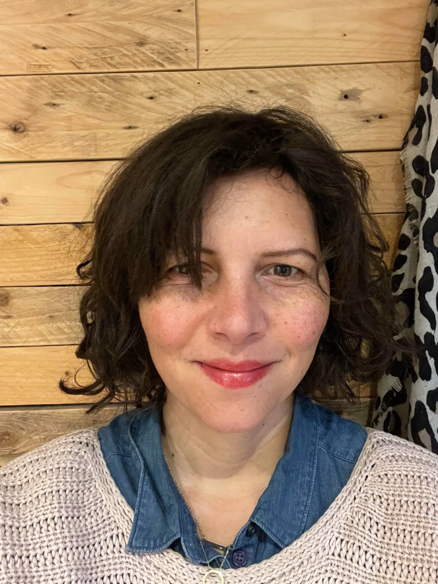 A woman with dark, curly hair and fair skin taking a selfie in front of a wooden background, wearing a blue denim shirt and a pink sweater.