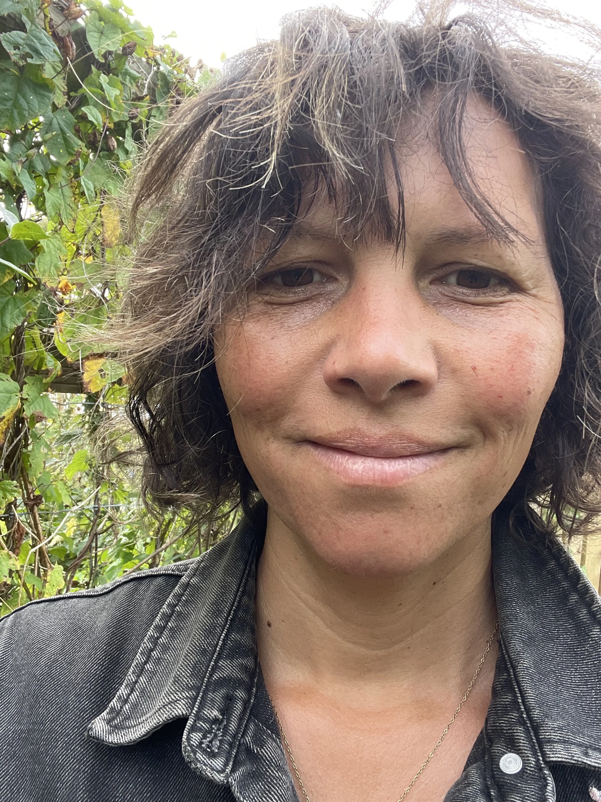A woman with short, wavy brown hair, slightly messy, smiling gently outdoors, with green foliage in the background, wearing a black denim jacket and a thin gold chain necklace.