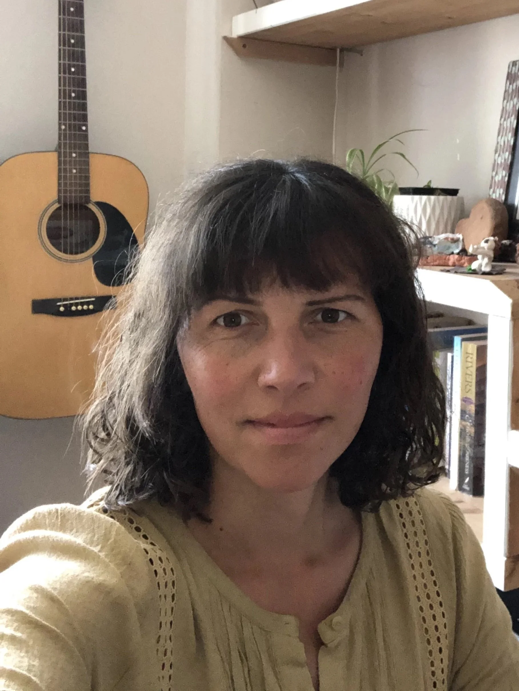 A woman taking a selfie in a room with a guitar hanging on the wall behind her and a bookshelf on the right side with various items and books.