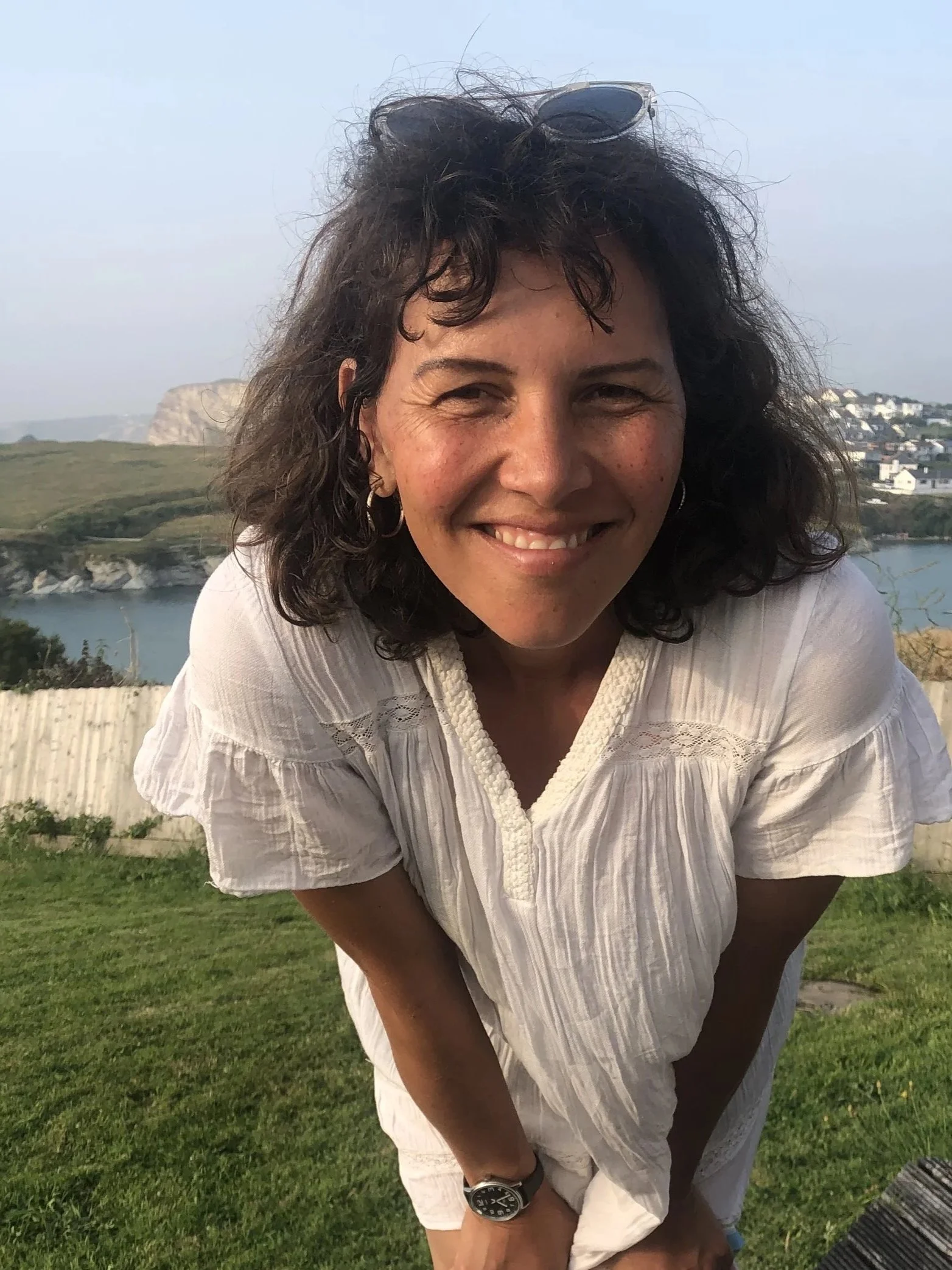 A woman with curly dark hair is smiling and leaning forward outdoors with a body of water, hills, and houses in the background.