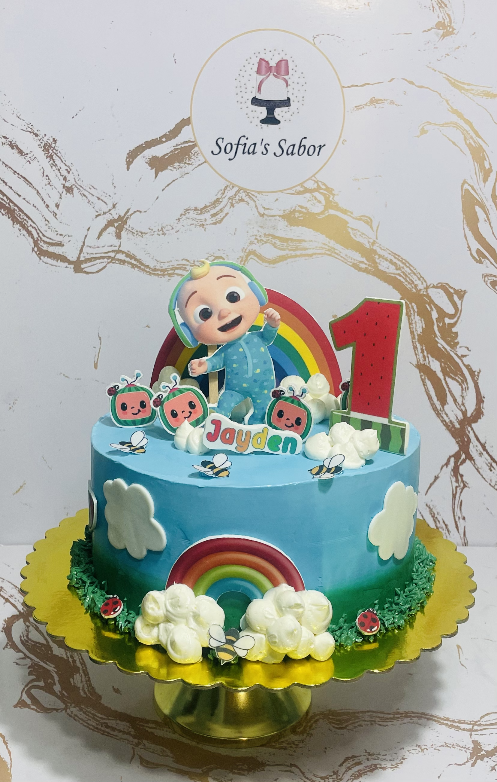 Pastel decorado para el primer cumpleaños de un niño, con tema de bebé sonriente, arcoíris, mariquitas, abejas y nubes en colores vivos. El nombre 'Jayden' y el número '1' están presentes. Fondo de diseño sofía.