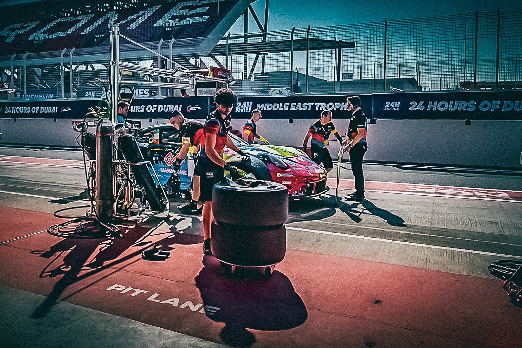 Echipa Willi Motorsport în plin pit-stop în timpul zilei la 24H Dubai, coordonând alimentarea și schimbul de pneuri pentru Porsche-ul GT3 în culori vibrante.