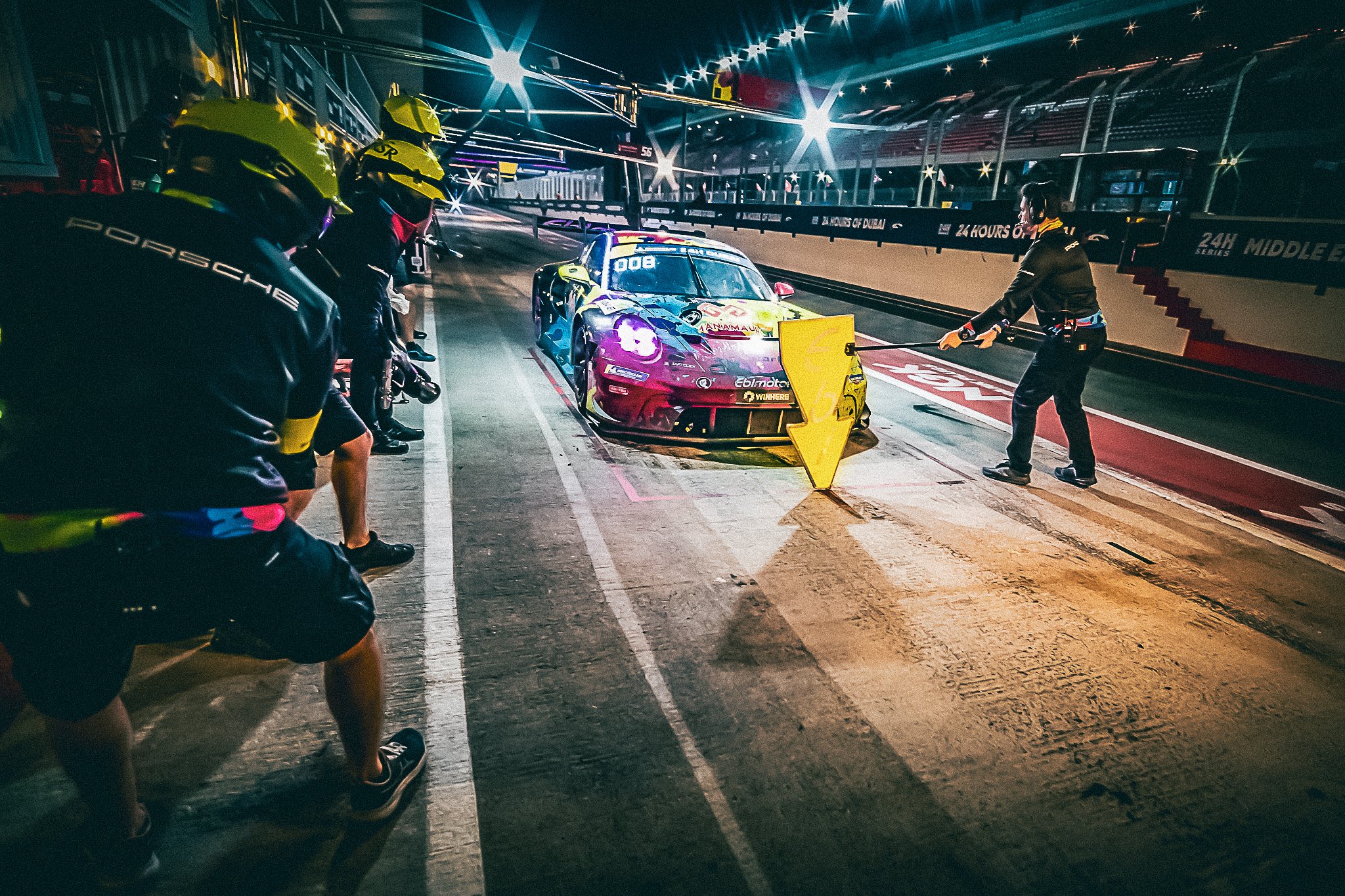 Momentul premergător pit-stop-ului la 24H Dubai, cu echipa Willi Motorsport pregătită să intervină pe Porsche-ul GT3 colorat, sub luminile nocturne ale paddock-ului.