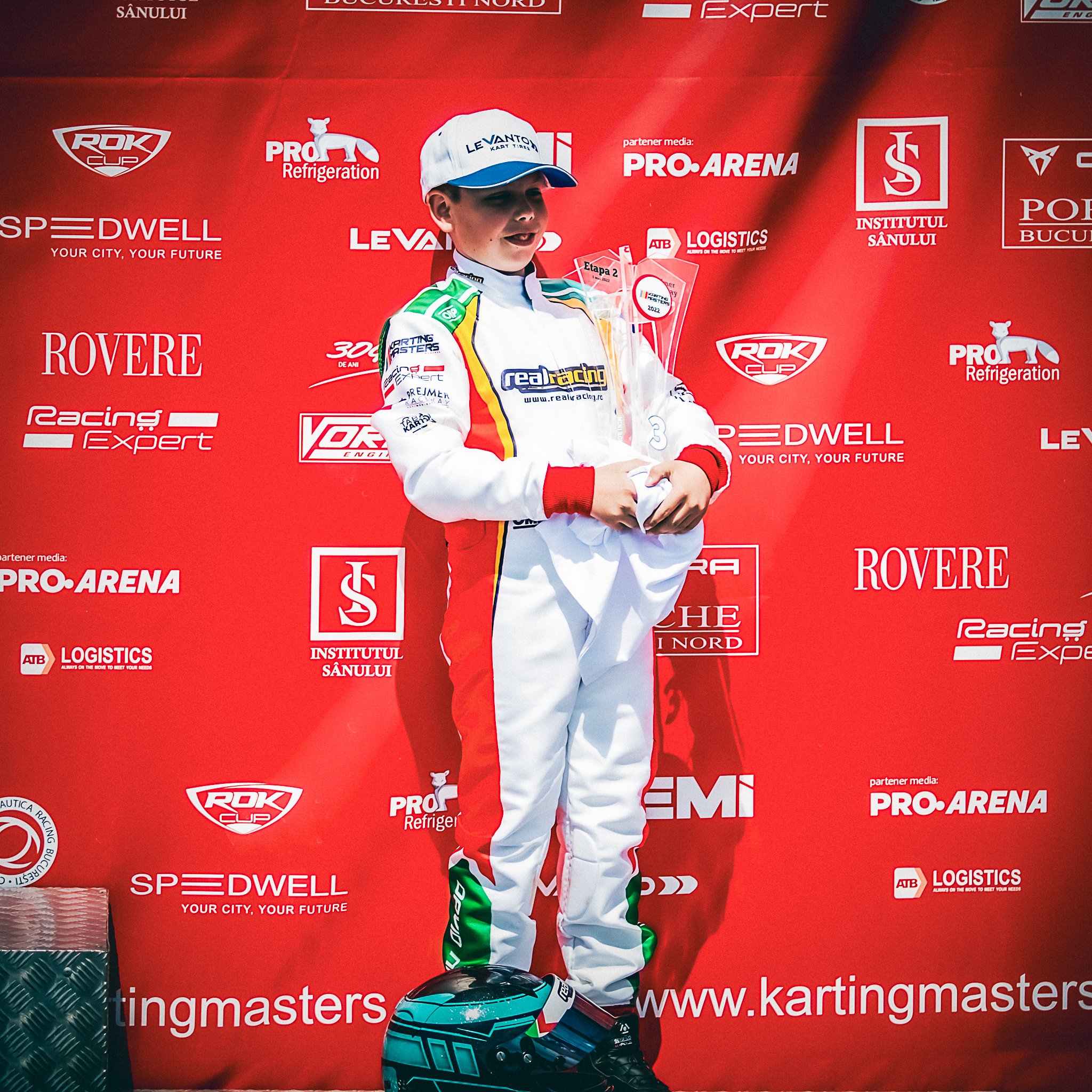 David Nicolaie pe podium, ținând trofeul obținut la o etapă Karting Masters ROK Cup, în combinezonul echipei Real Racing, în fața panoului oficial al competiției.