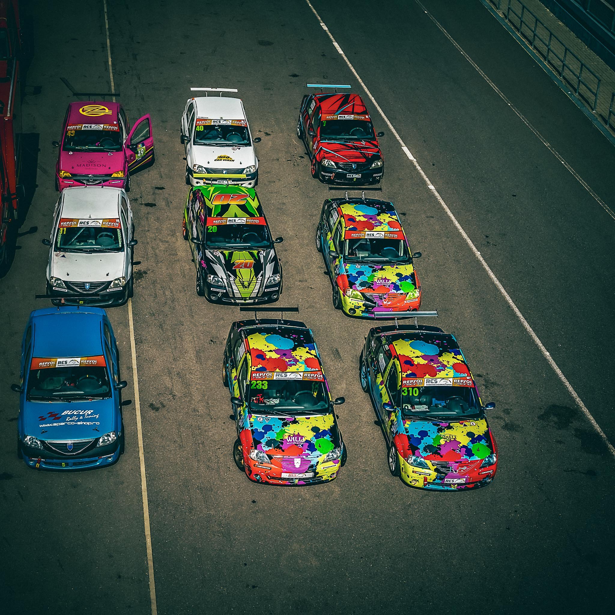 Vedere de sus cu mașinile din Logan Cup aliniate în paddock, înainte de cursă, inclusiv mașina #233 pilotată de David Nicolaie, cu design colorat și vibrant.