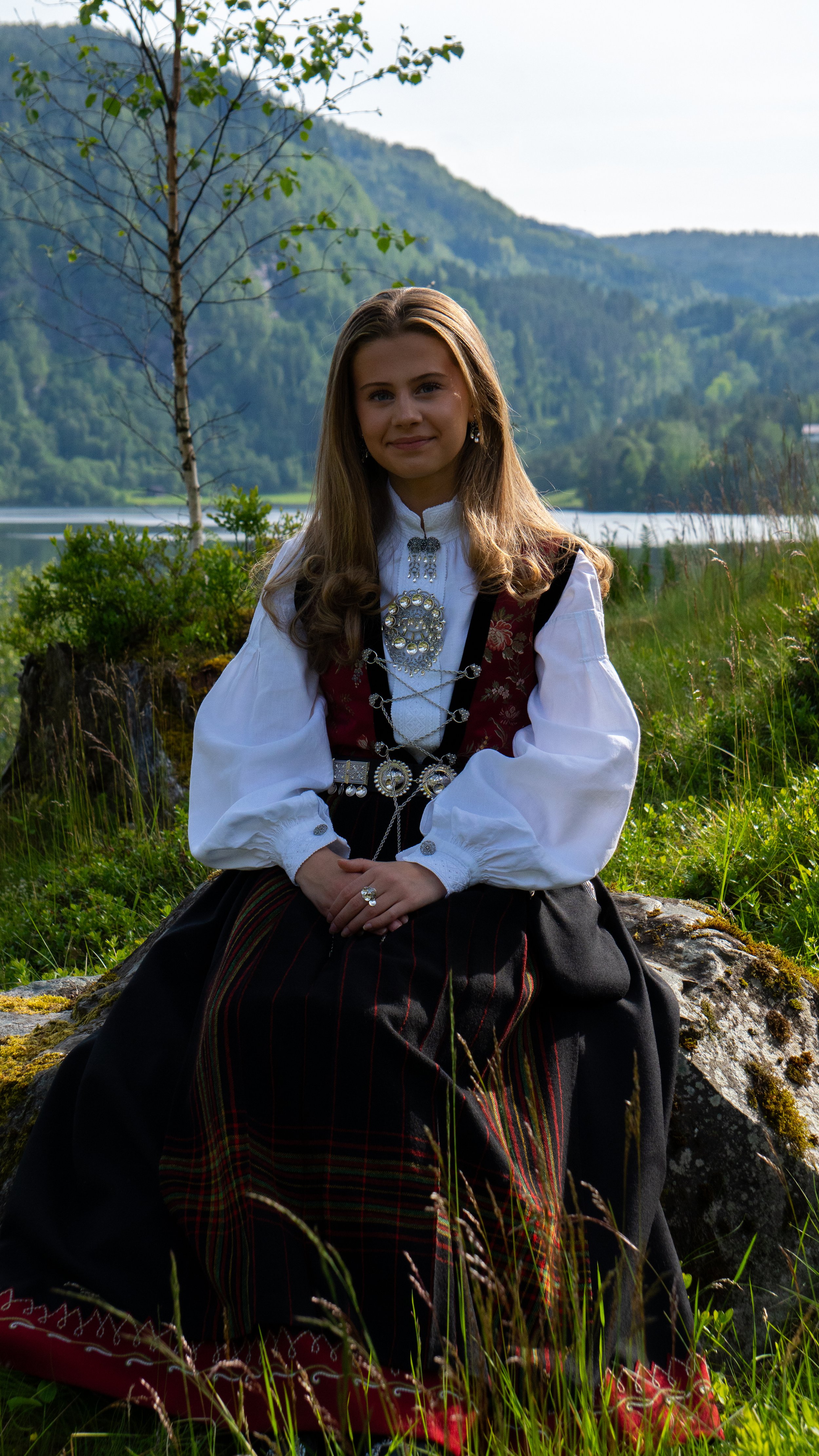 A young woman in traditional Norwegian folk dress sitting outdoors on a rock, with a lake, green trees, and mountains in the background.