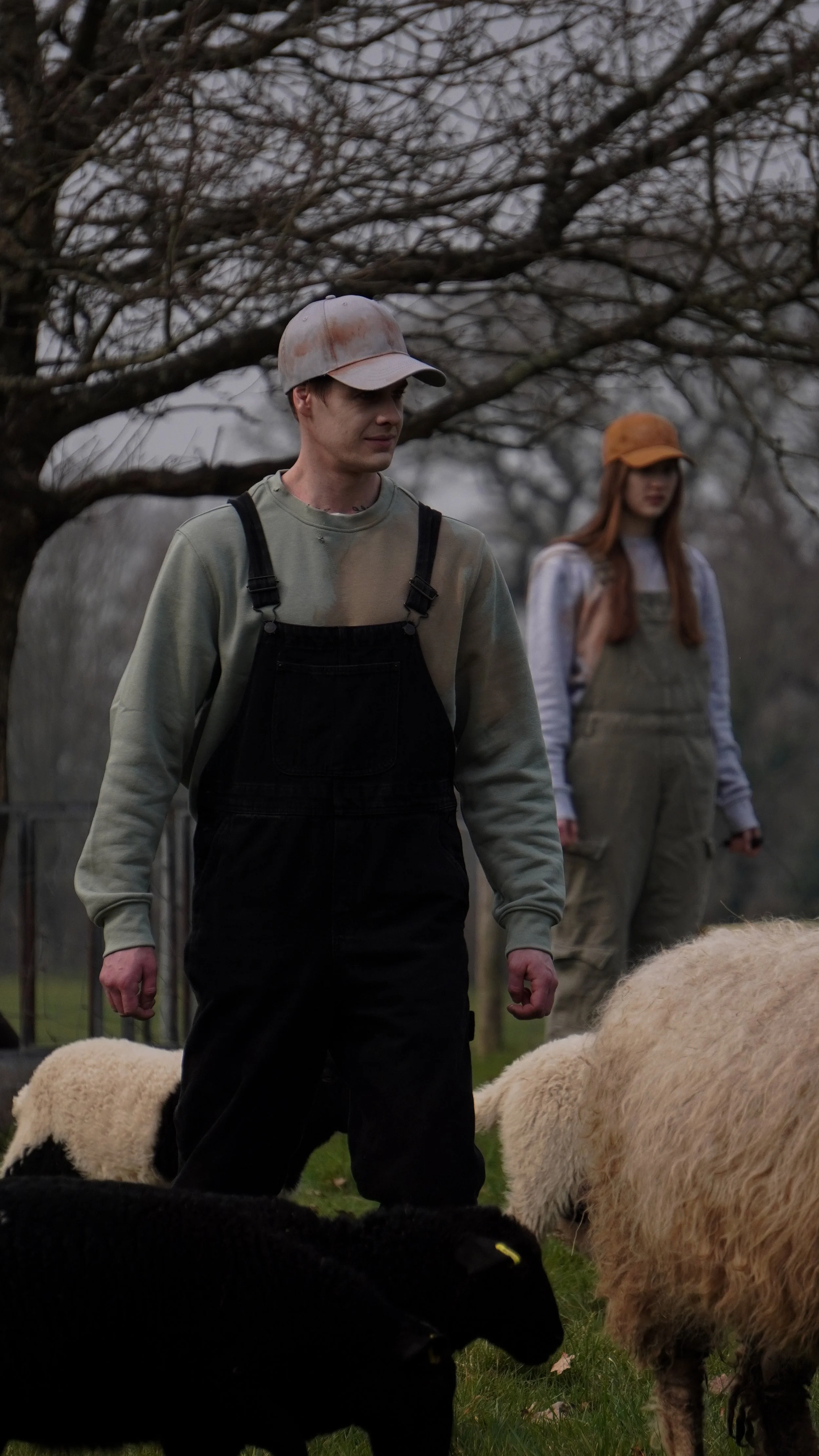 A young man and woman stand outdoors among sheep, with trees in the background. The man wears a beige cap, a gray sweatshirt, and black overalls, while the woman wears an orange hat, a gray shirt, and tan overalls. They appear to be on a farm or rural setting.