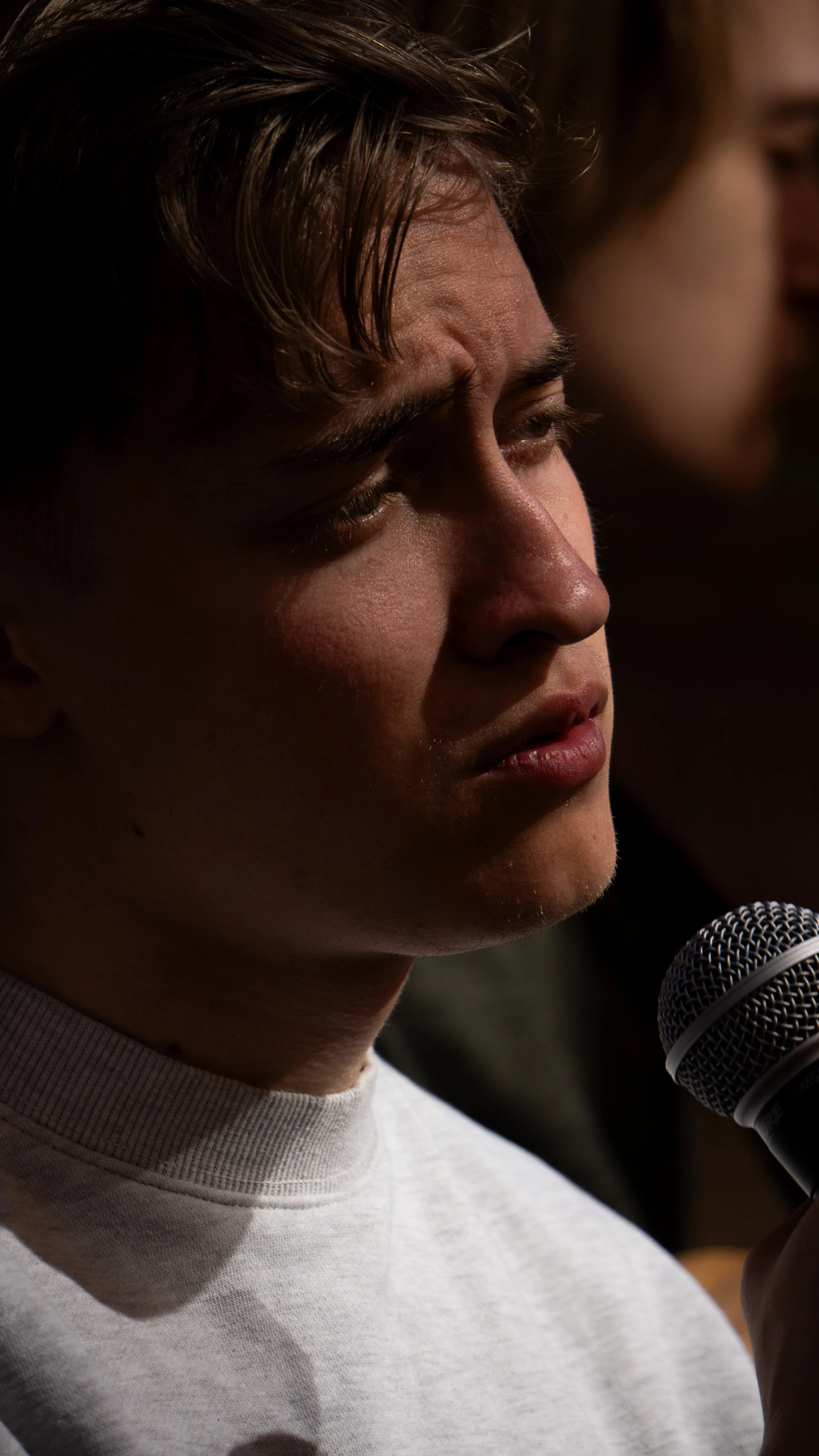 Young man with short hair and light skin holding a microphone, appearing to speak or sing, with a woman slightly out of focus in the background.