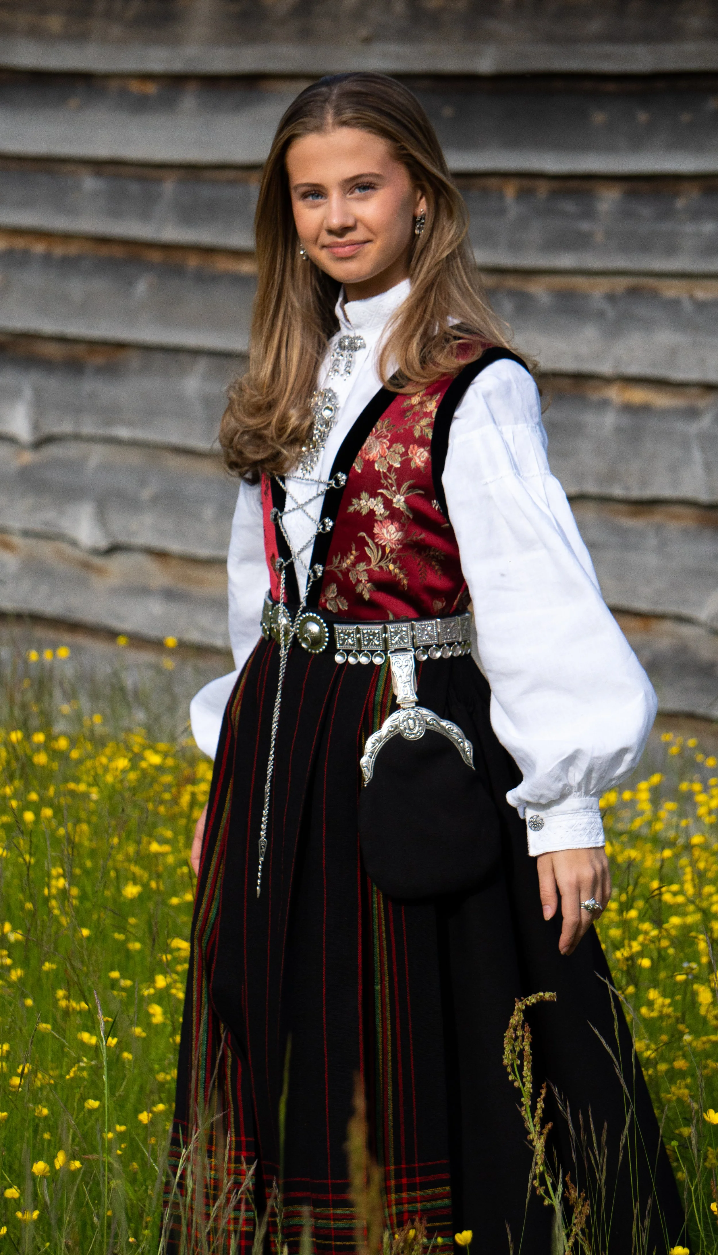 A young woman wearing traditional Norwegian folk costume, standing in a field of yellow flowers with a wooden building in the background.
