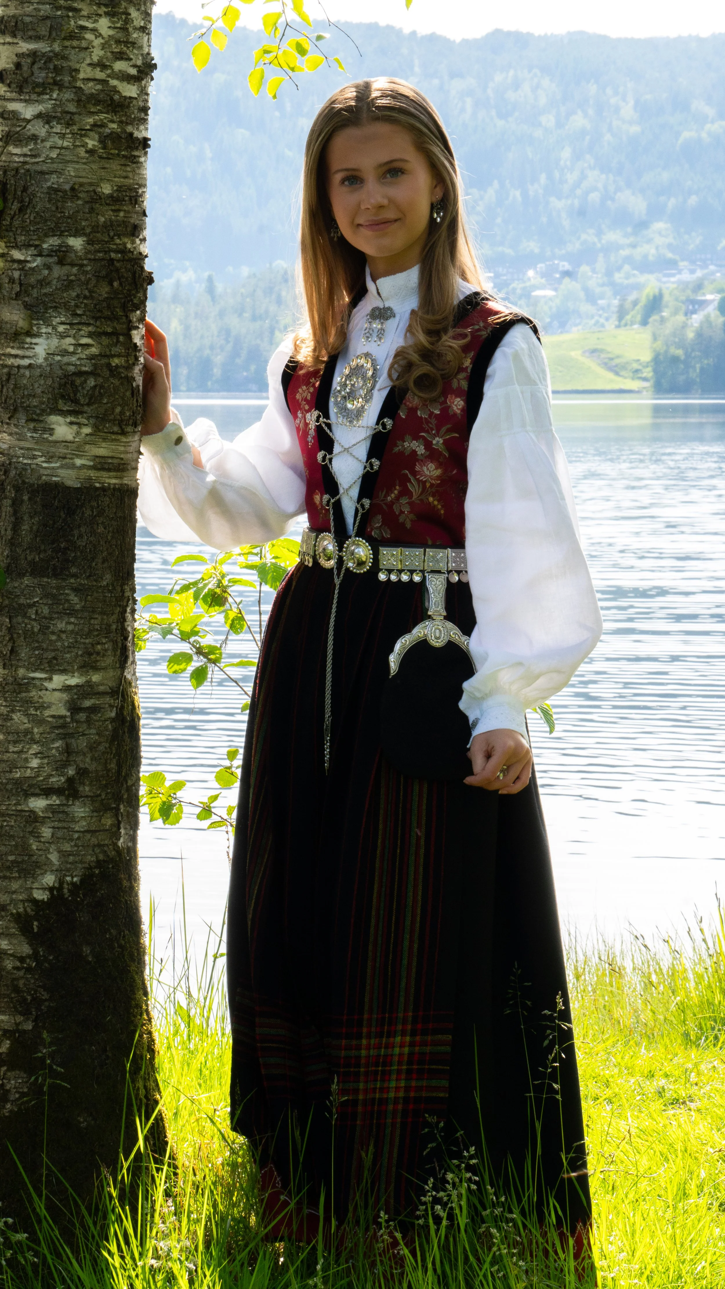 Young woman outdoors by a lake, wearing traditional Norwegian folk costume with jewelry, standing near a tree with a mountain and lake in the background.