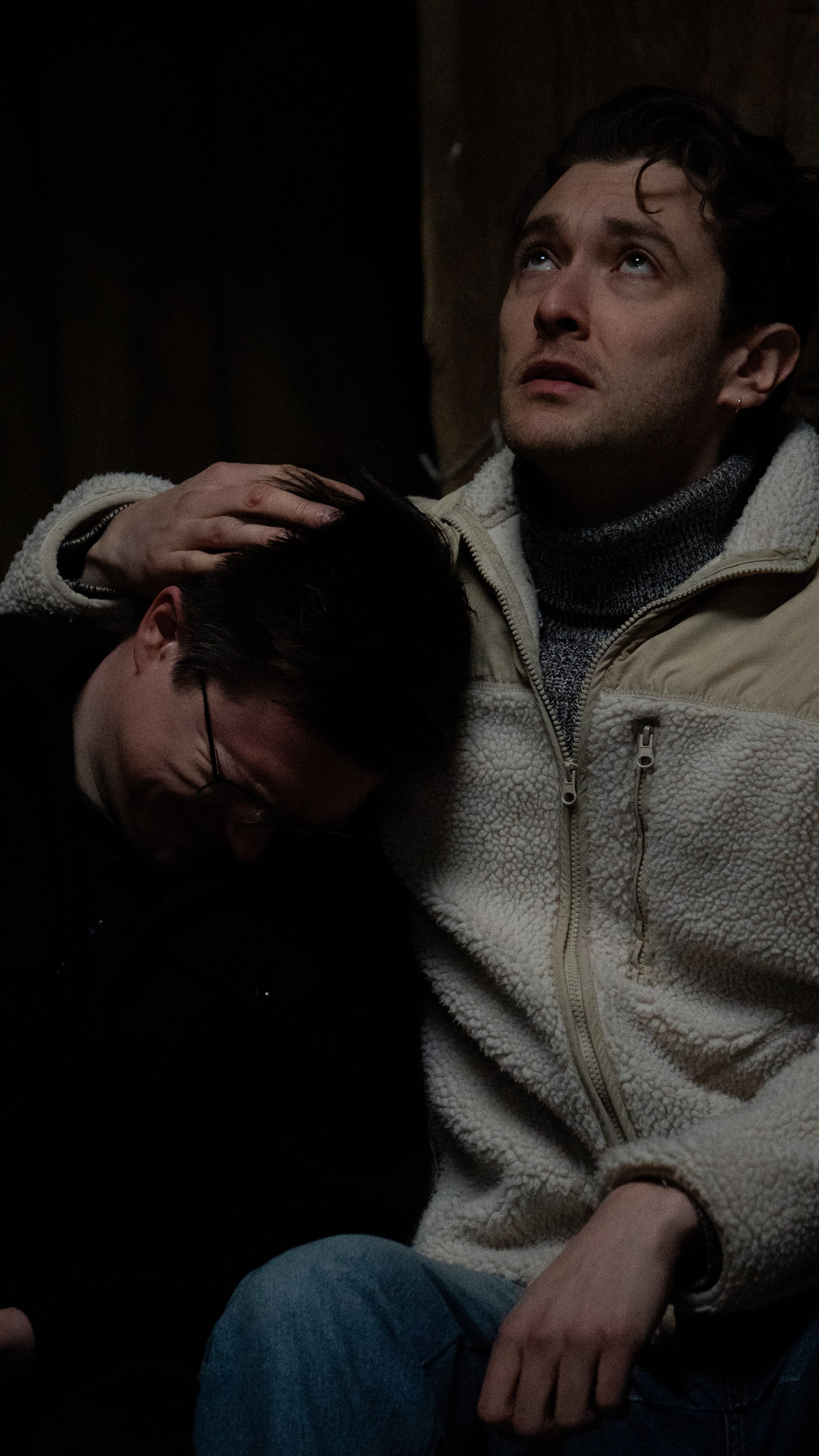 A man with dark hair and light skin, wearing a beige fleece jacket and gray turtleneck, is sitting with a woman with glasses and dark hair who is leaning on his chest. He is gently placing his hand on her head, and she appears to be resting her head against his shoulder in a comforting gesture.