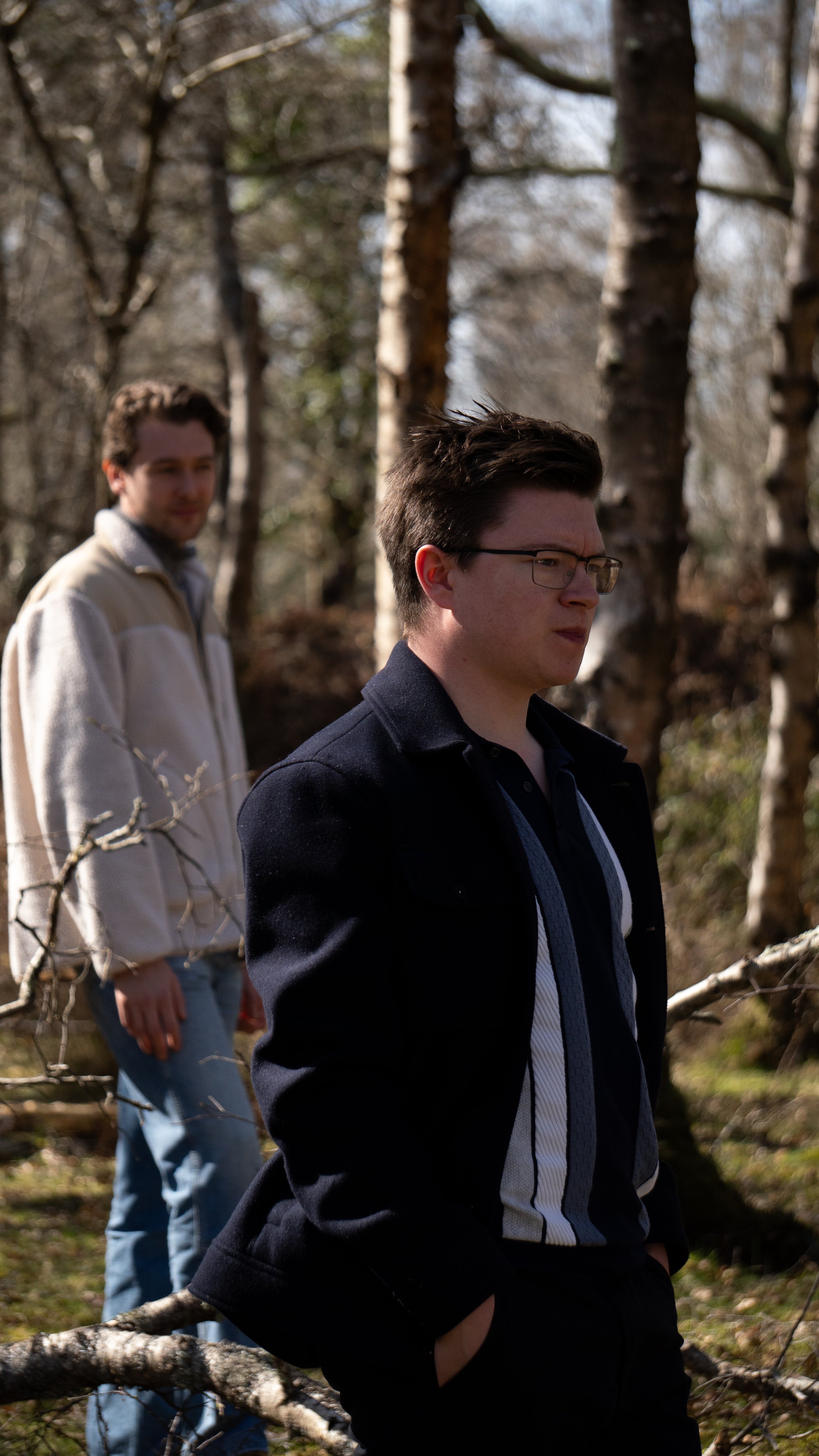 Two young men standing outdoors among trees in a forest, with one in front wearing glasses and a dark jacket and the other in the background wearing a light jacket and jeans.
