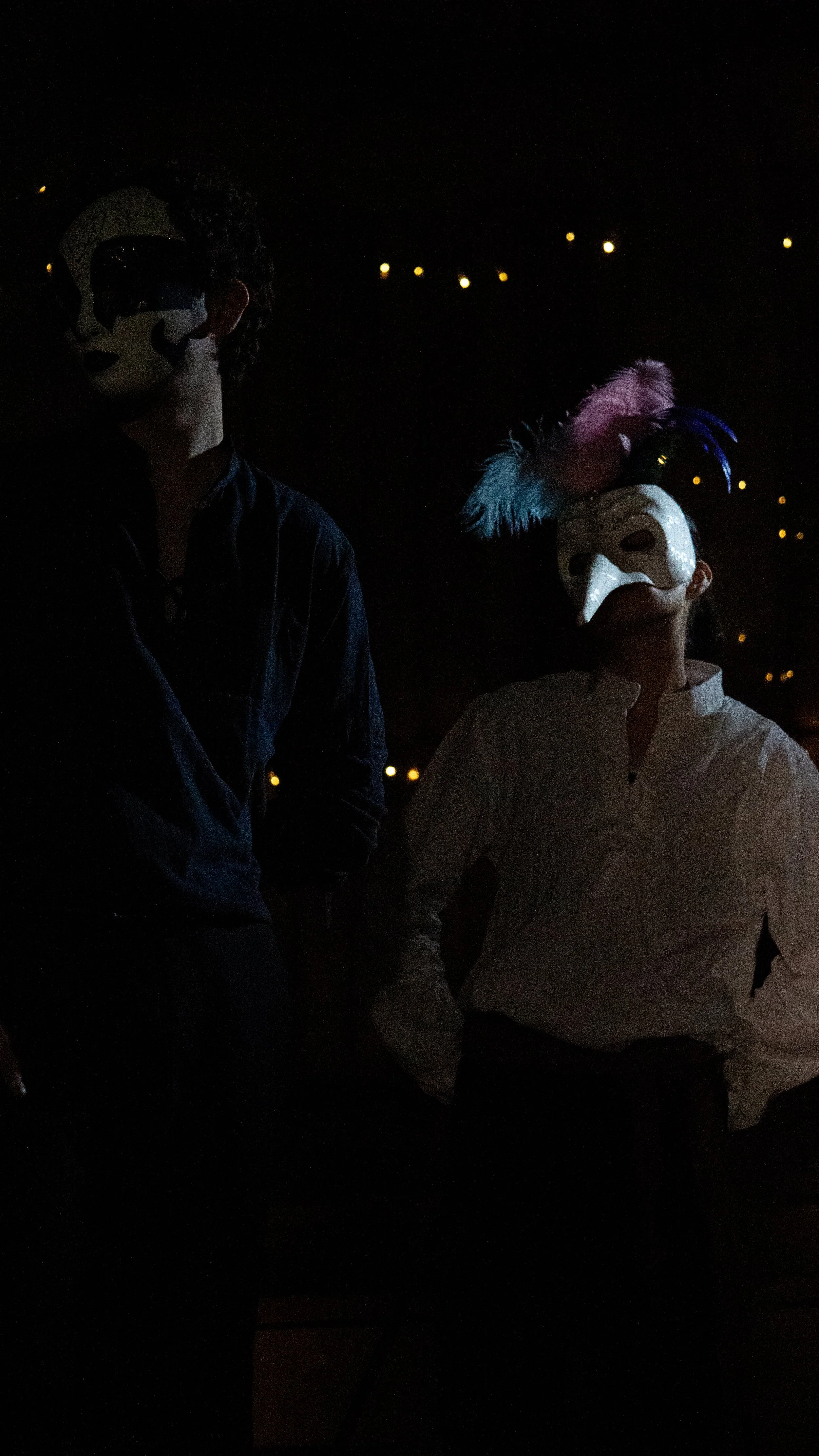 Two people wearing decorative masks with feathers and face paint, standing in a dark environment with small yellow lights in the background.