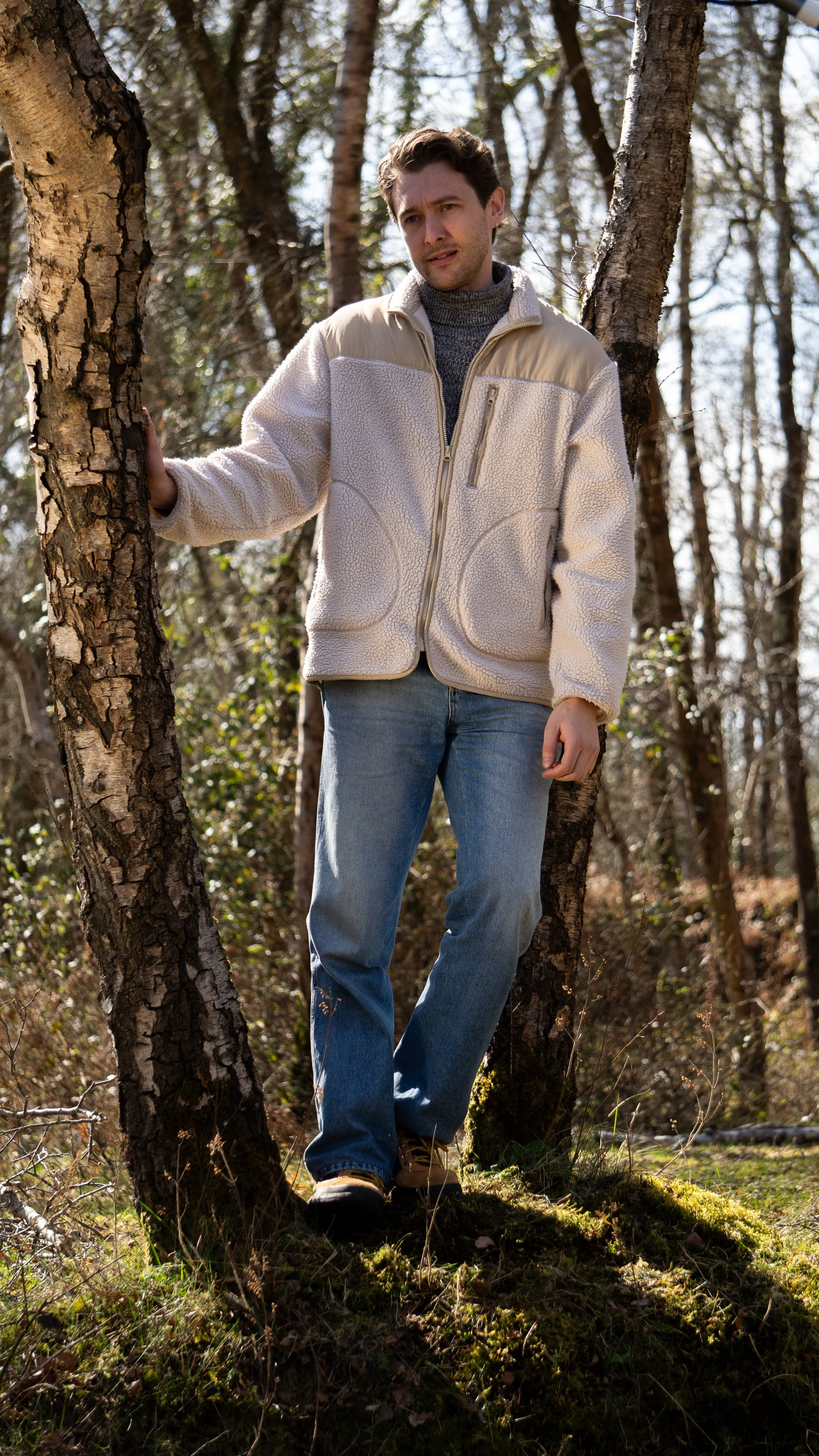 A man standing among trees in a forest during daytime, wearing a beige and cream fleece jacket, a gray turtleneck, blue jeans, and tan boots.