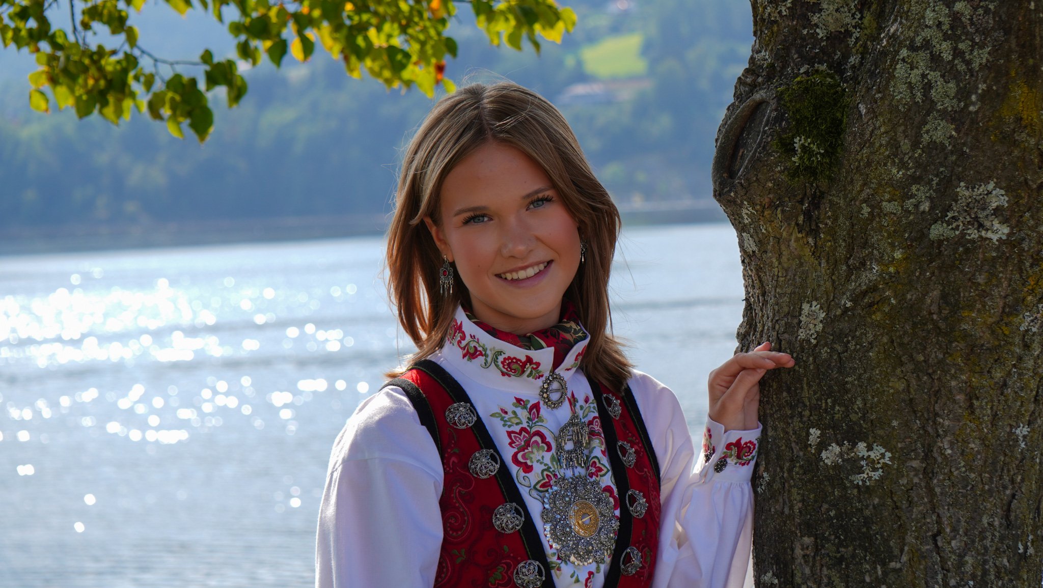 A young woman dressed in traditional folk attire, standing outdoors near a body of water, smiling and holding onto a tree trunk.