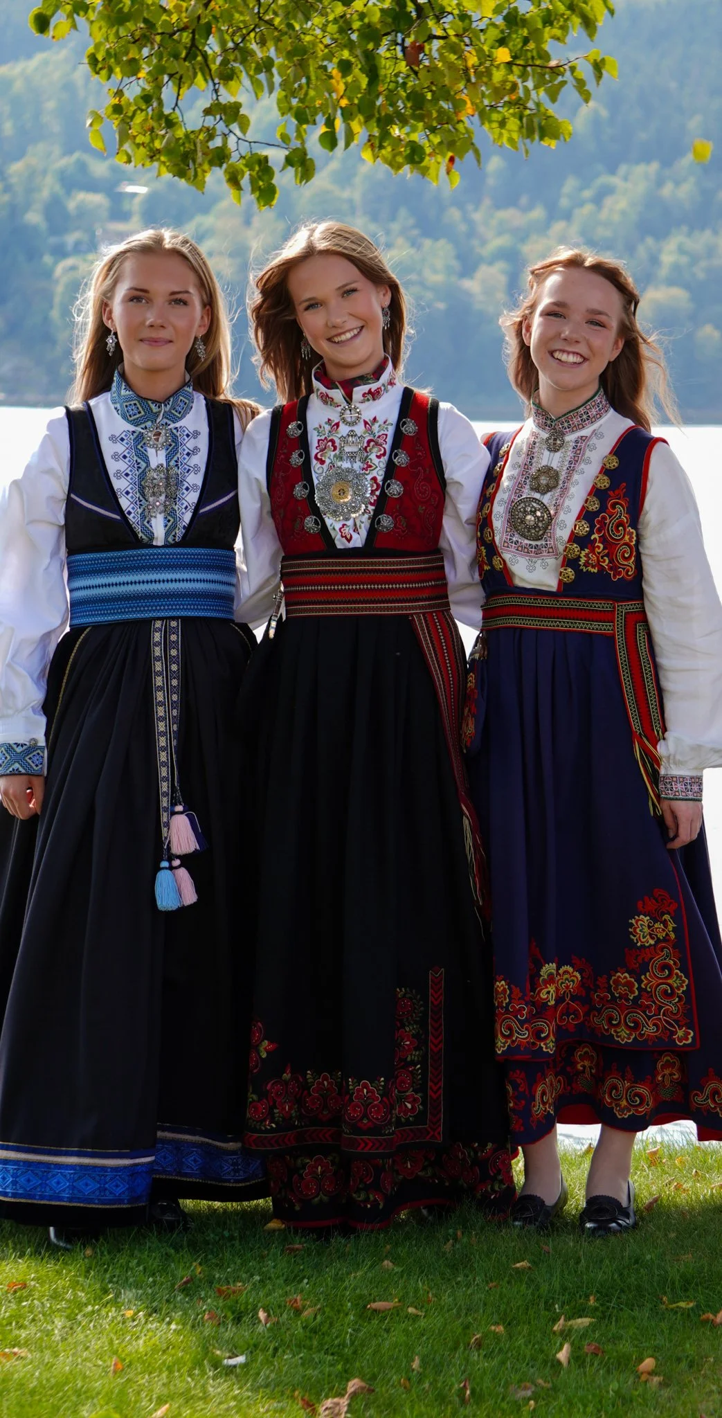 Three young women in traditional Norwegian folk costumes stand outdoors near a lake, with trees and mountains in the background.