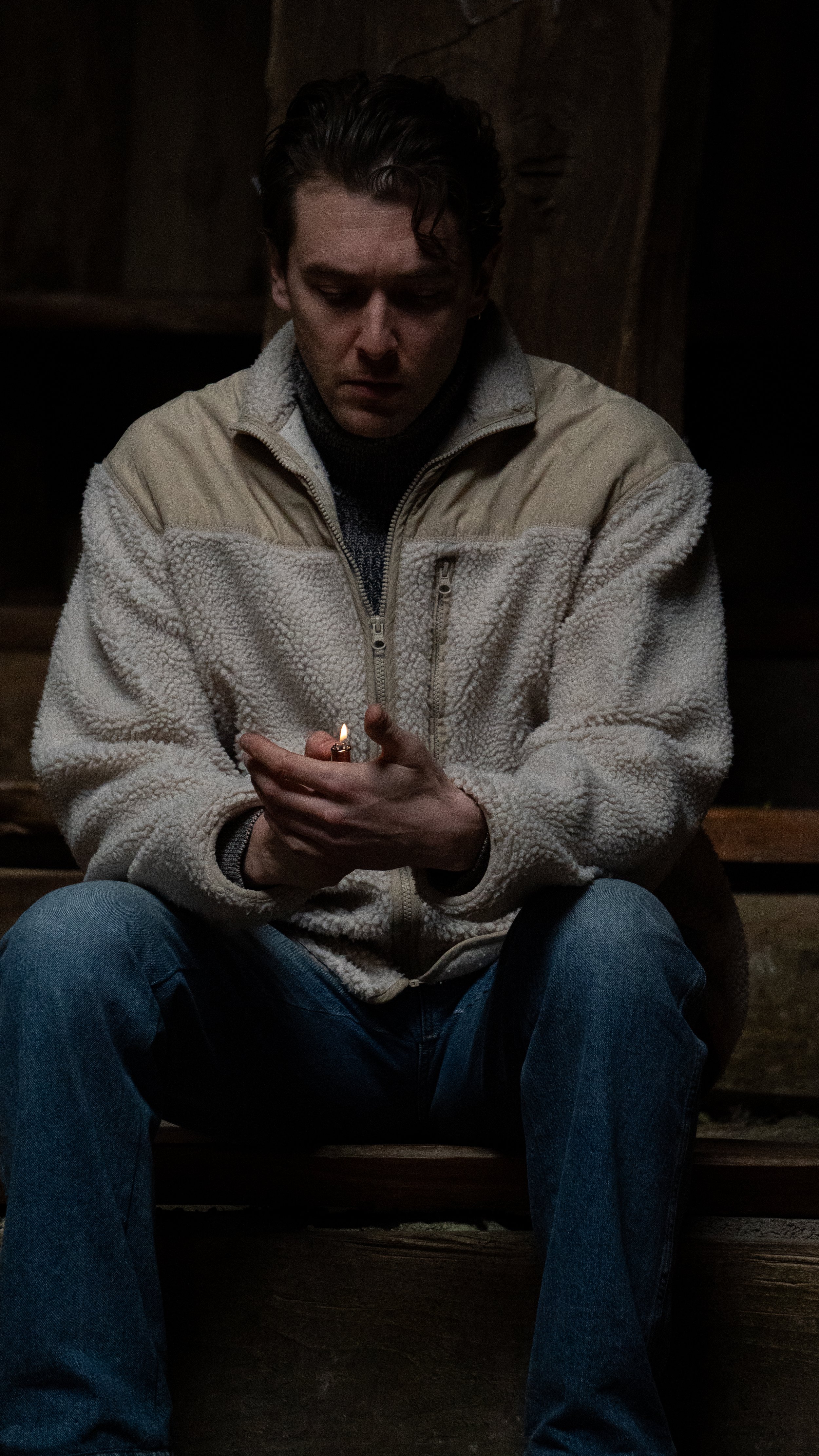 A man sitting outdoors in a dark setting, holding a lighter with a small flame, wearing a light-colored fleece jacket and jeans.