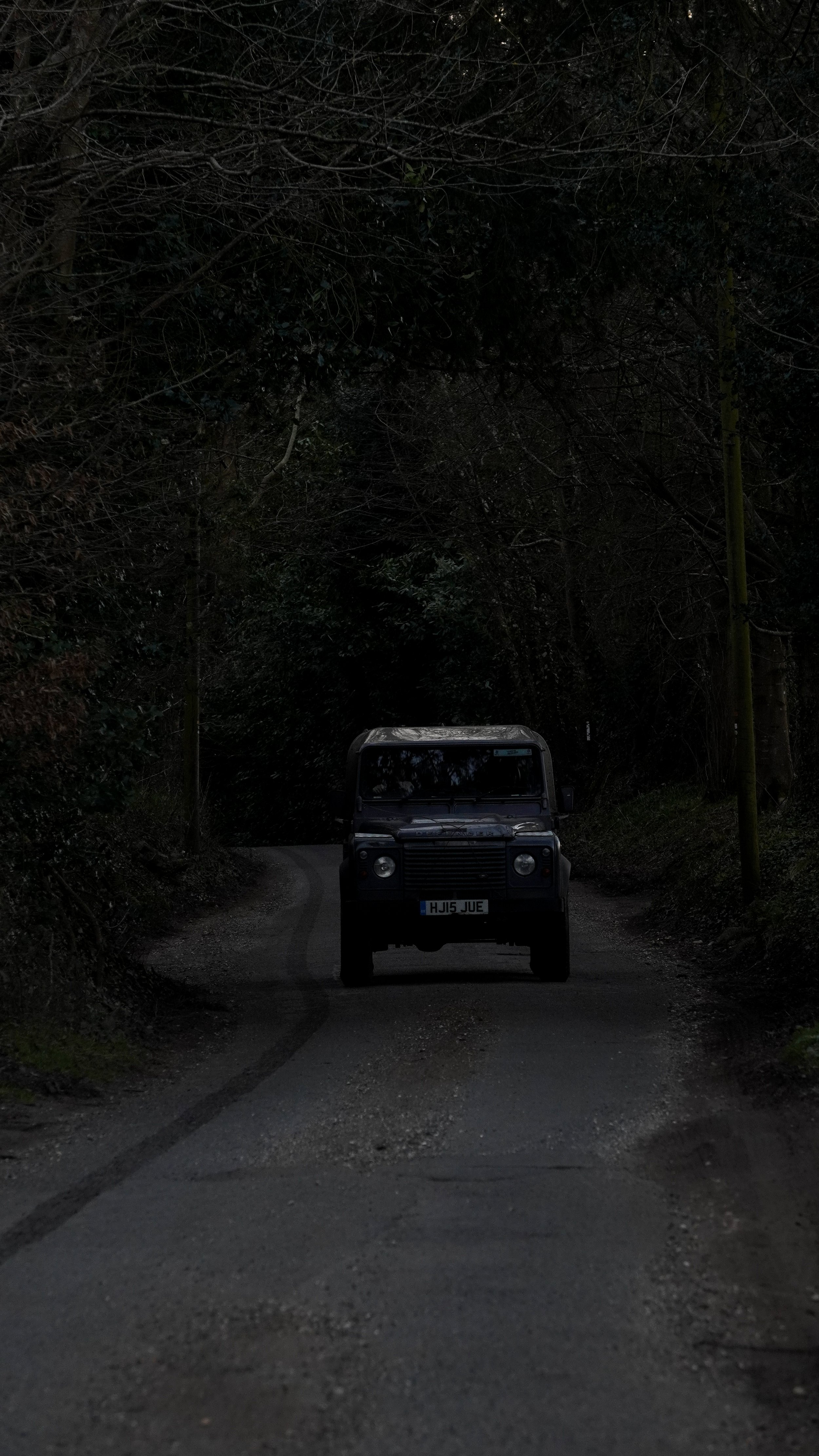 A Land Rover Defender driving on a narrow, winding dirt road surrounded by dense trees and foliage.