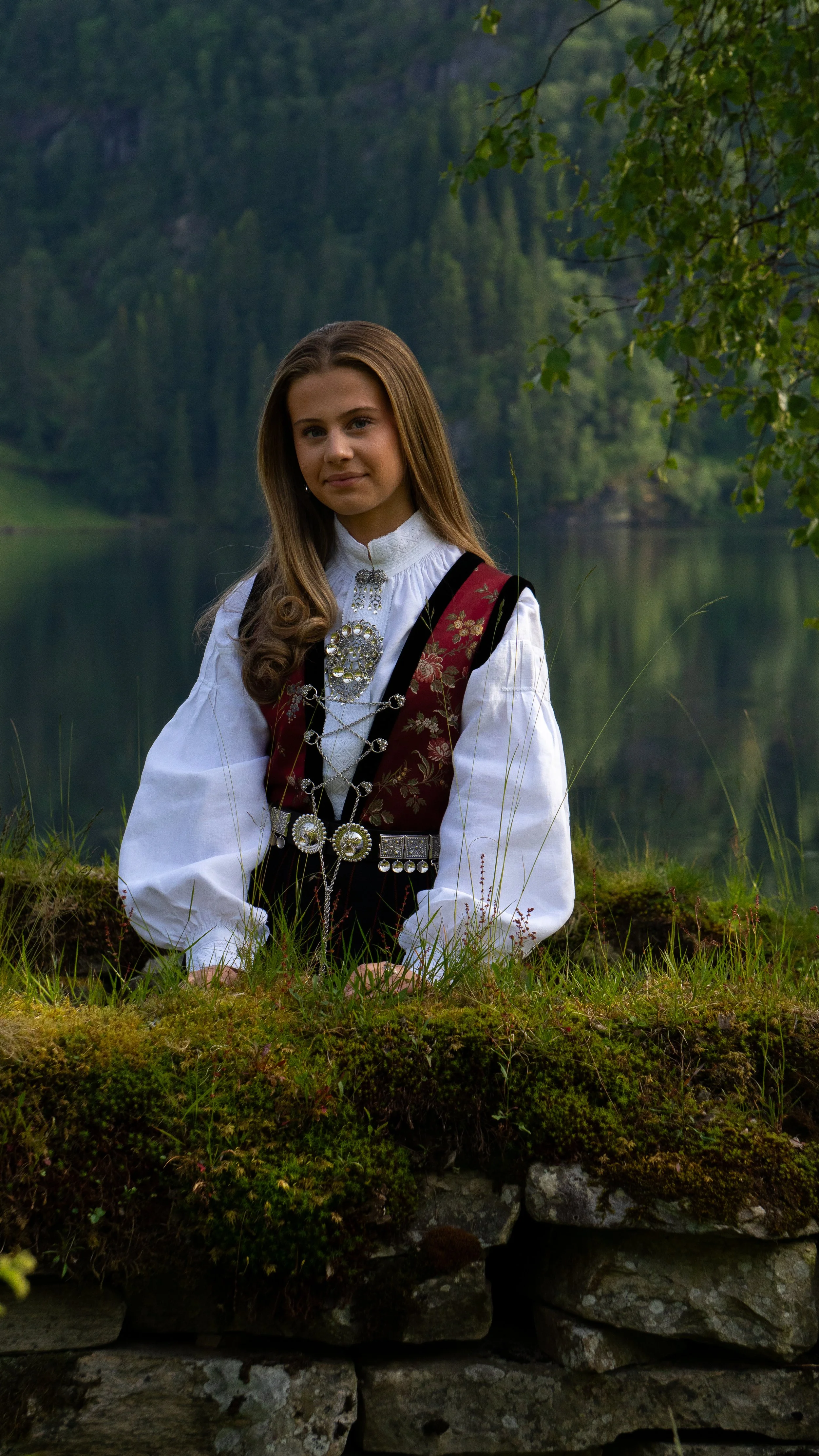 A young girl in traditional European folk costume sitting outdoors near a lake with lush green trees and mountains in the background.