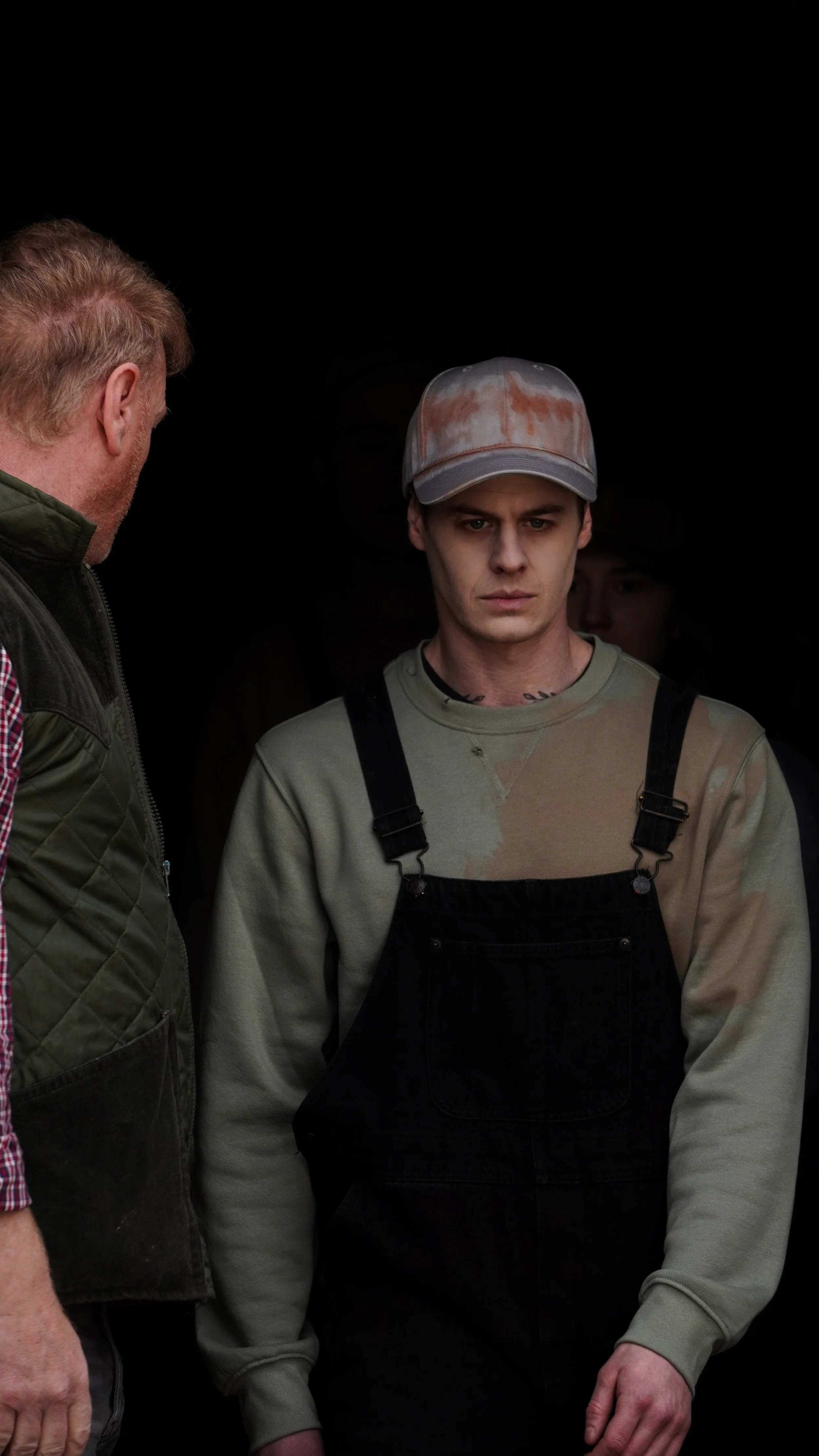 A young man wearing a dirty baseball cap and overalls looks serious while talking to an older man wearing a green jacket in a dark setting.