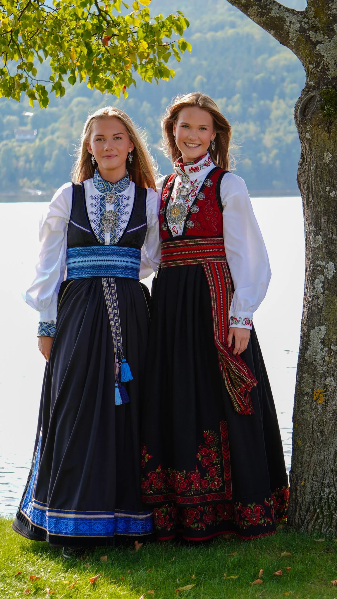 Two women in traditional Norwegian folk costumes standing outdoors near a tree by a river, with a forested hillside in the background.