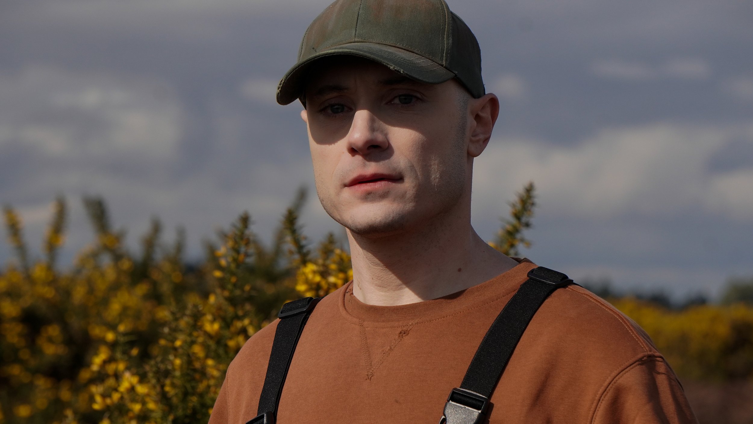 A young man wearing a green baseball cap and a brown shirt with black suspenders, standing outdoors with yellow shrubs and a cloudy sky in the background.