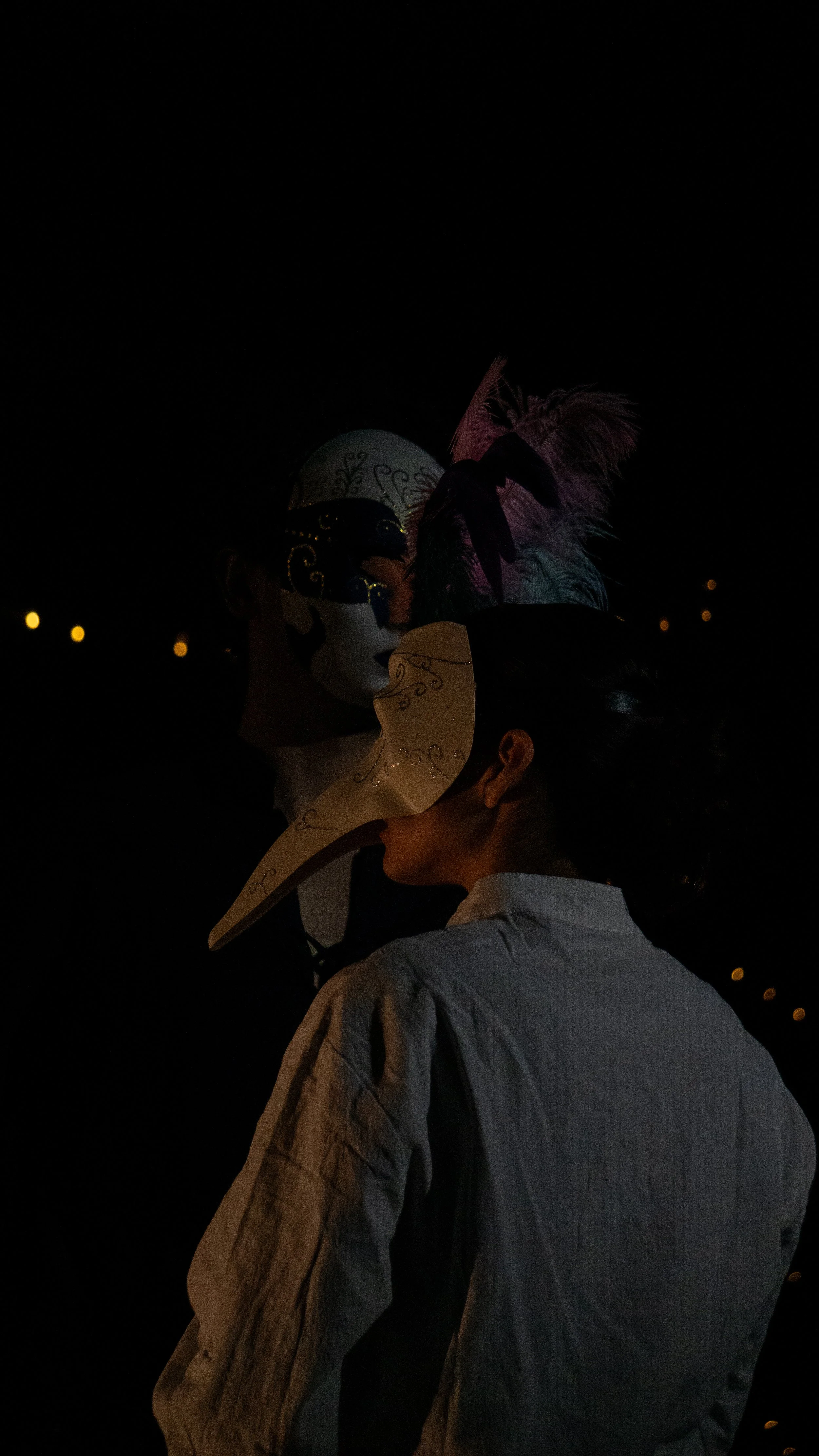 Person wearing a white shirt and a decorative mask with a large, ornate feathered headpiece, standing near another person with a painted face and a fan-shaped mask in a dark environment with small lights in the background.