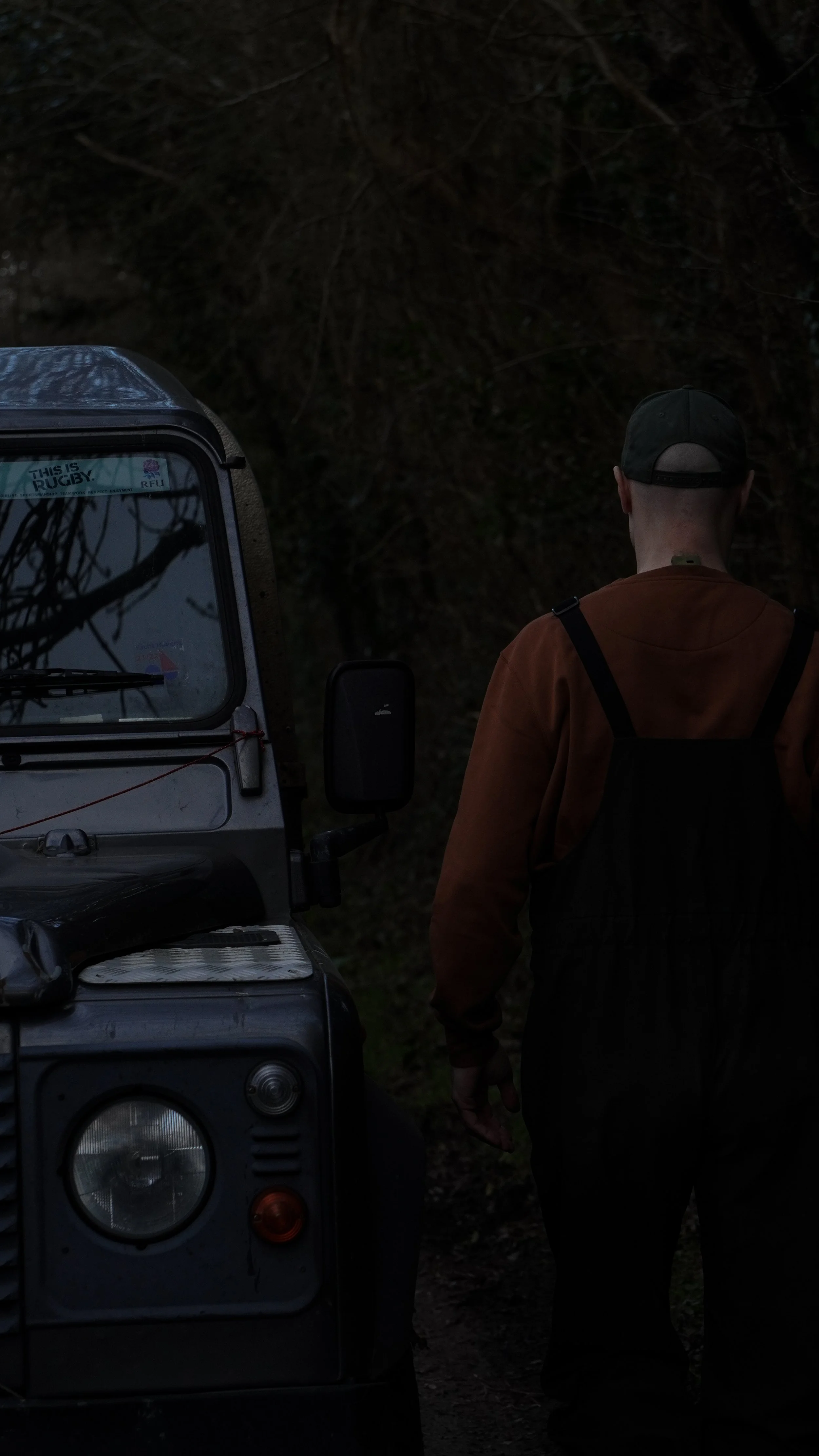 A person standing next to a vehicle at dusk or dawn, with trees and dark sky in the background.