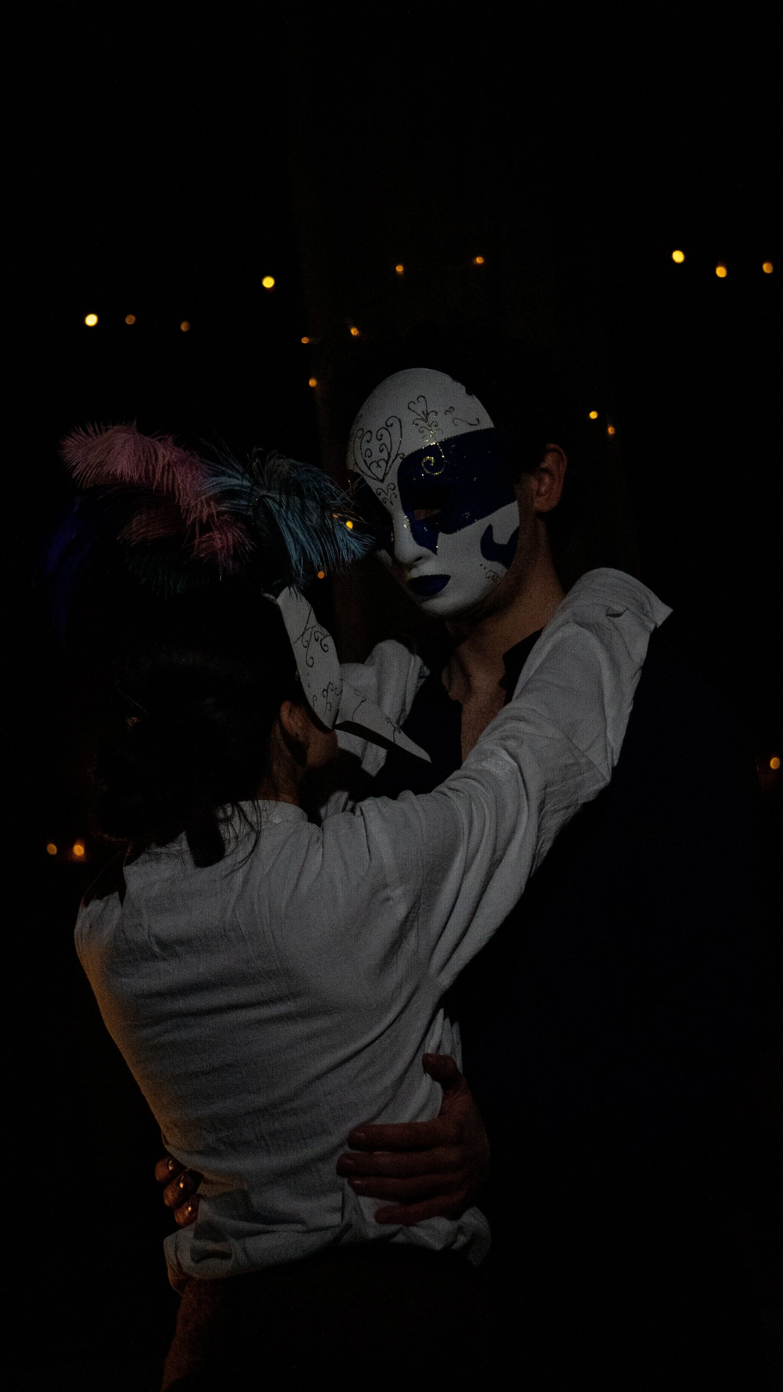 Two people in cosplay masks are embracing in a dark setting with small orange lights in the background.