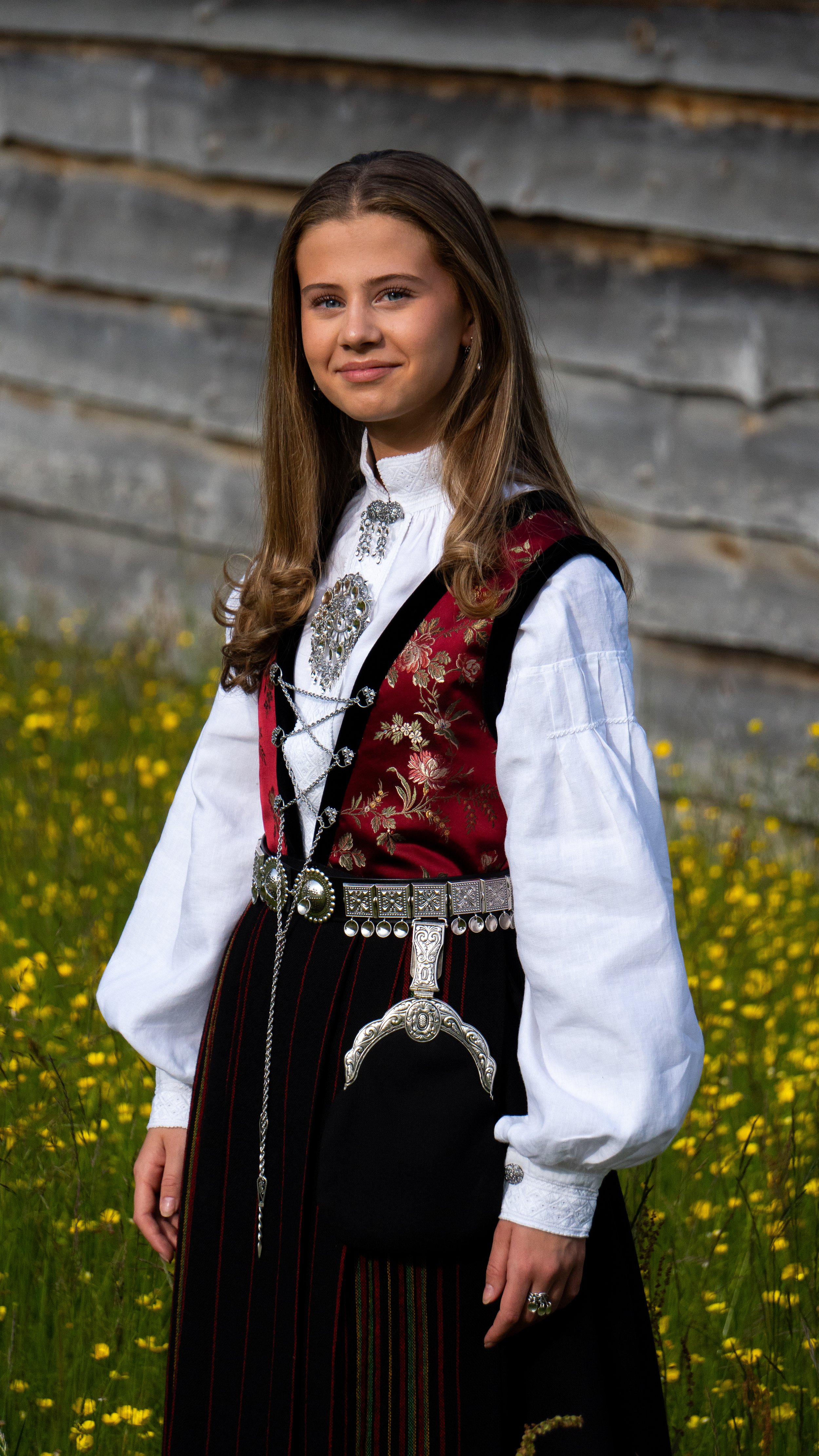 A young woman in traditional Norwegian attire standing in a field of yellow flowers with a rustic wooden fence in the background.