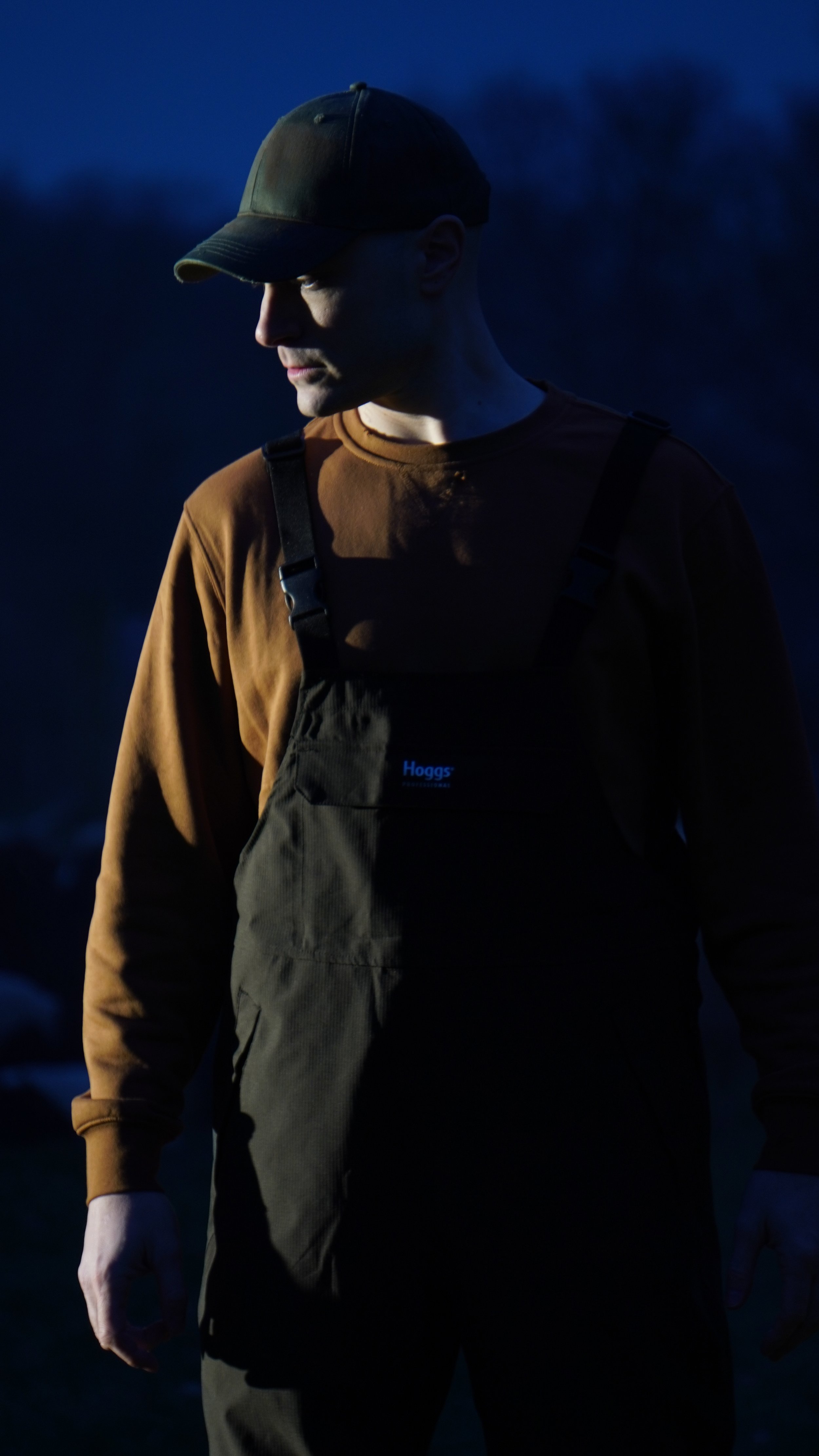 A man wearing a brown long-sleeve shirt, black overalls, and a dark cap, standing outdoors at dusk or dawn with a dark, cloudy sky in the background.