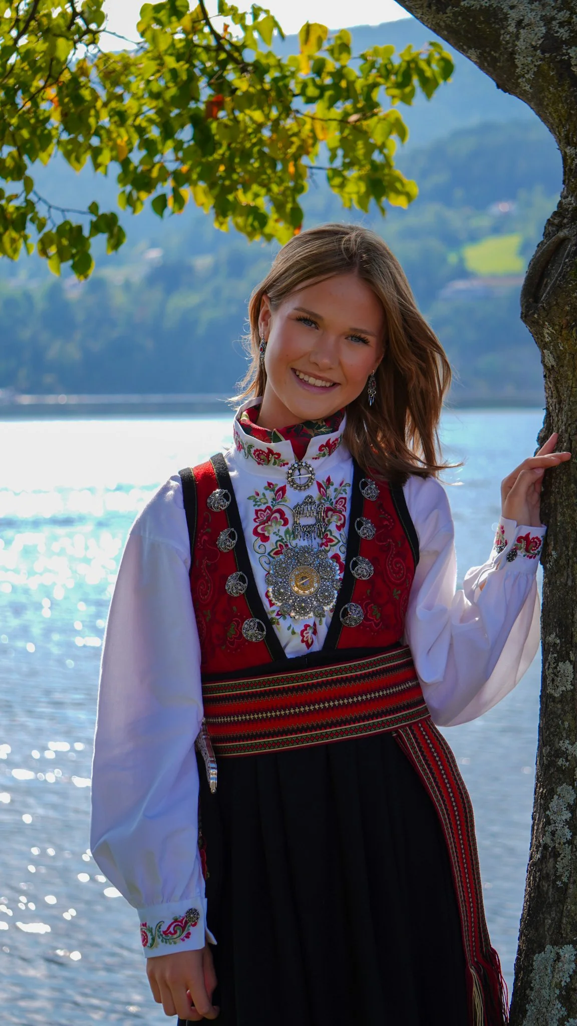 A smiling young woman in traditional Norwegian folk costume standing beside a tree by a lake with mountains in the background.