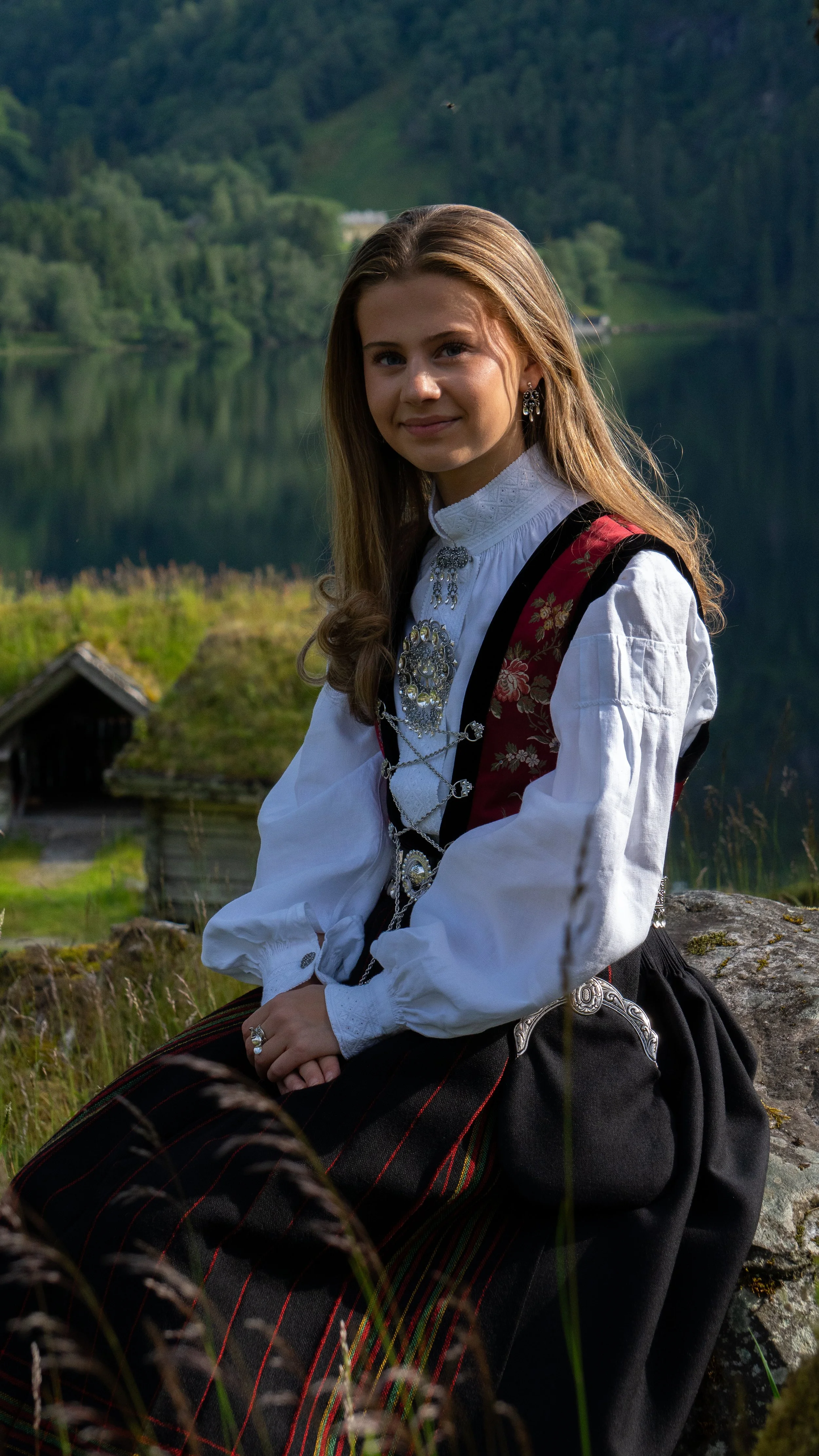 Young woman in traditional Norwegian bunad attire sitting outdoors near a lake with forested mountains in the background.