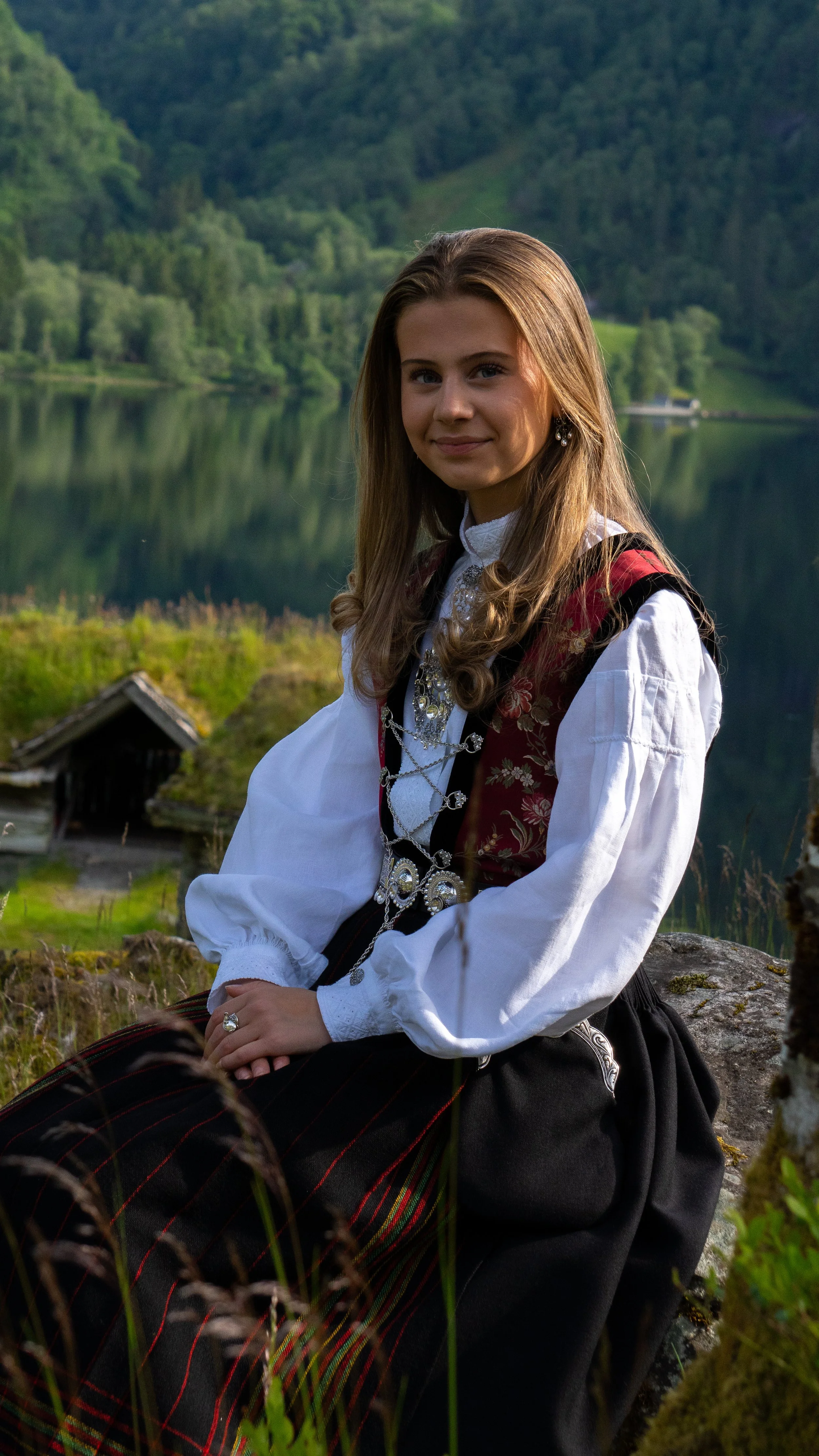 A young woman in traditional Norwegian dress sitting outdoors near a lake with green forested hills in the background.