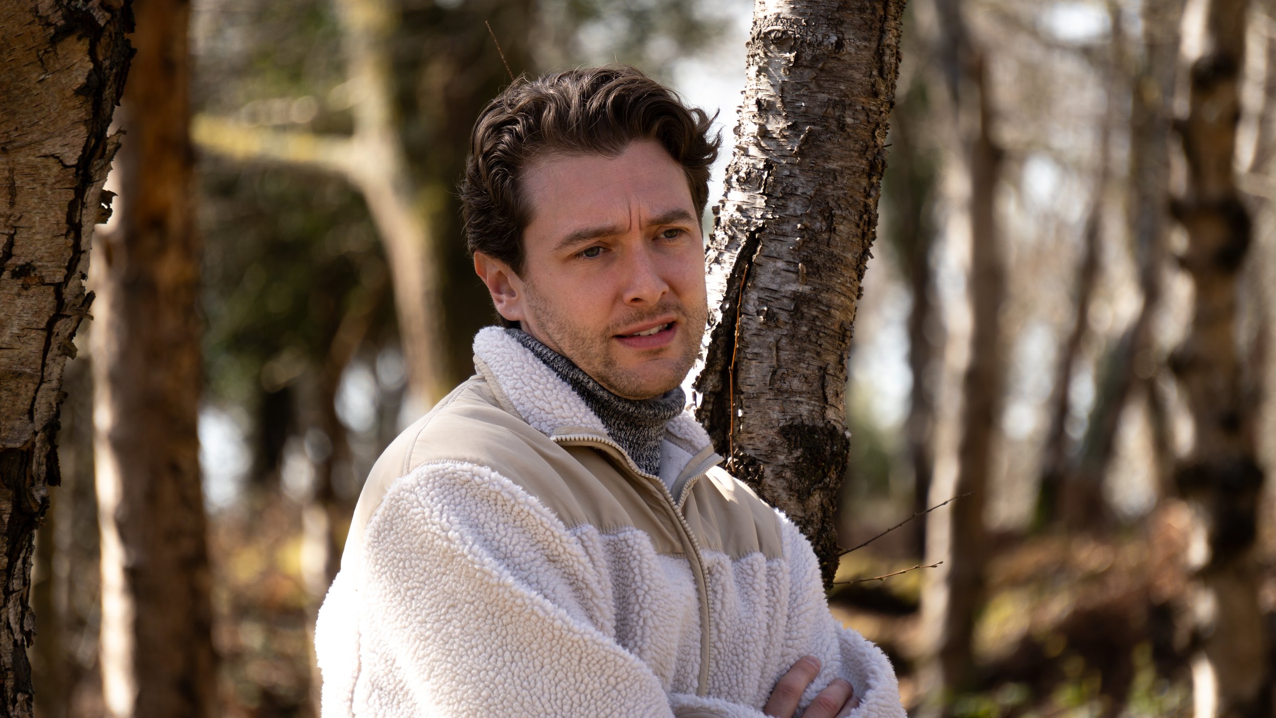 A man with dark hair and light skin standing outdoors among trees, wearing a beige and white fleece jacket and a gray turtleneck, looking to the side with a concerned expression.