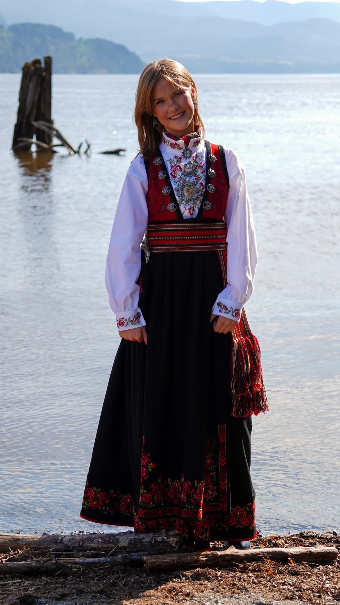 A young woman in traditional Norwegian clothing standing by a body of water with hills in the background.