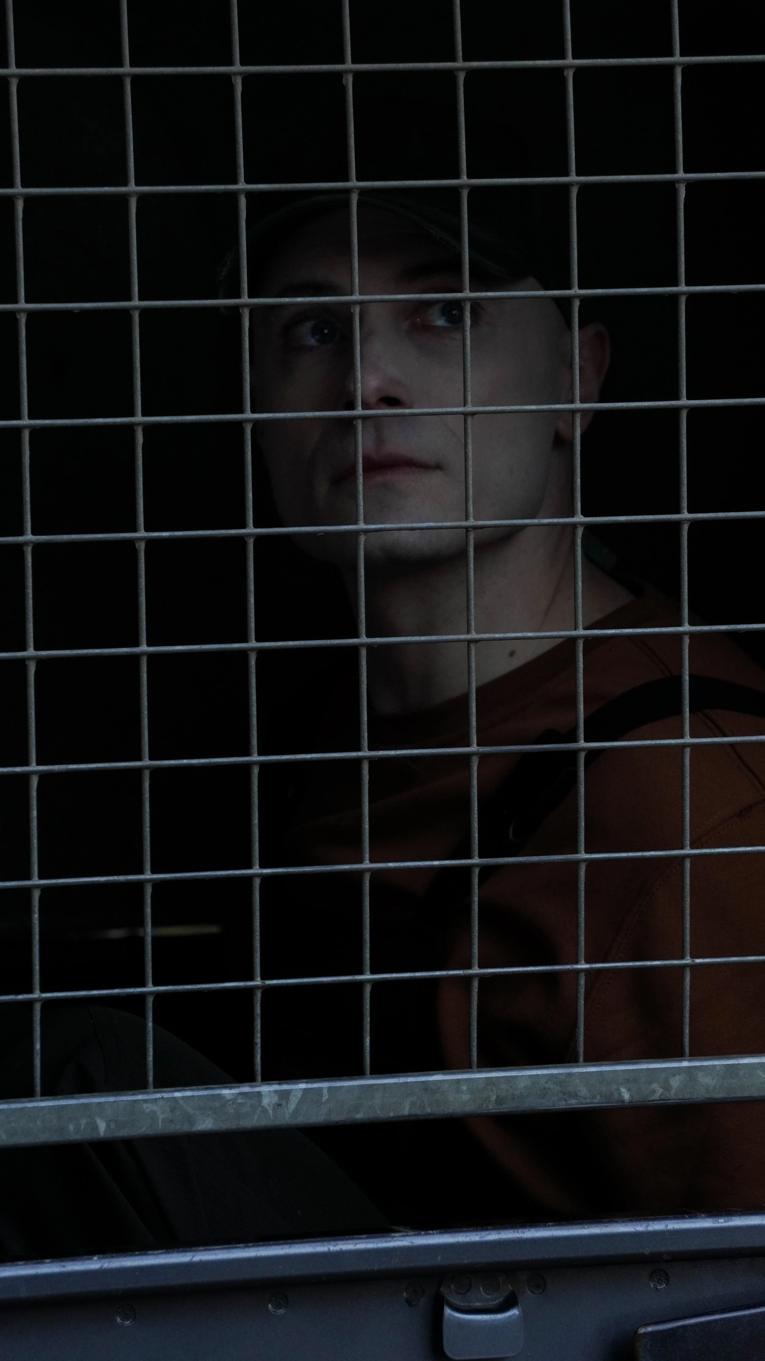 A man looking out from behind a metal grid, with a dark background, wearing an orange and black jacket.