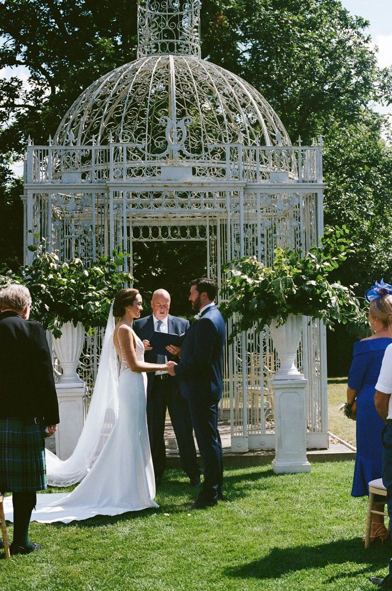 A wedding ceremony outdoors with a bride and groom exchanging vows in front of an ornate white gazebo, surrounded by guests.