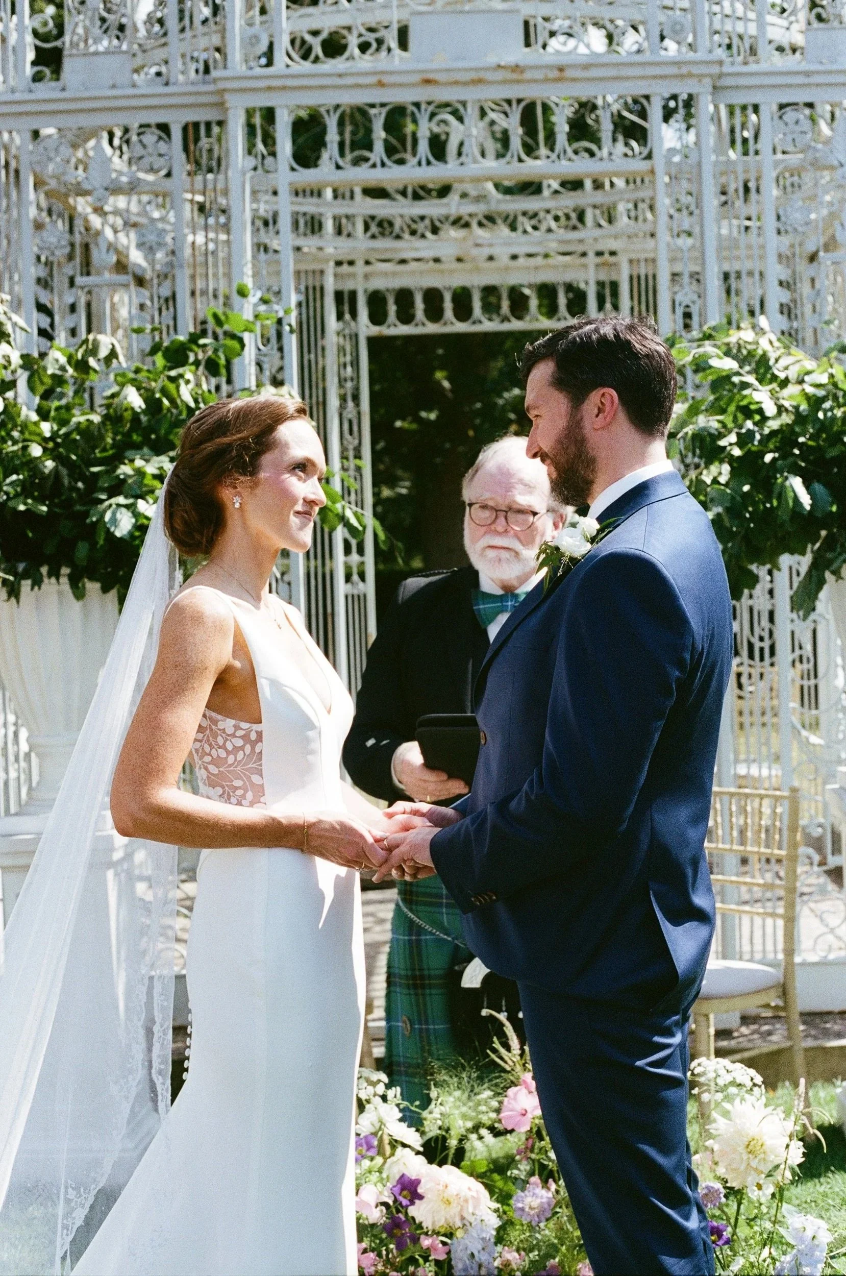 A bride and groom exchange vows at an outdoor wedding ceremony, holding hands and looking into each other's eyes, with an officiant standing behind them, surrounded by greenery and floral decorations.