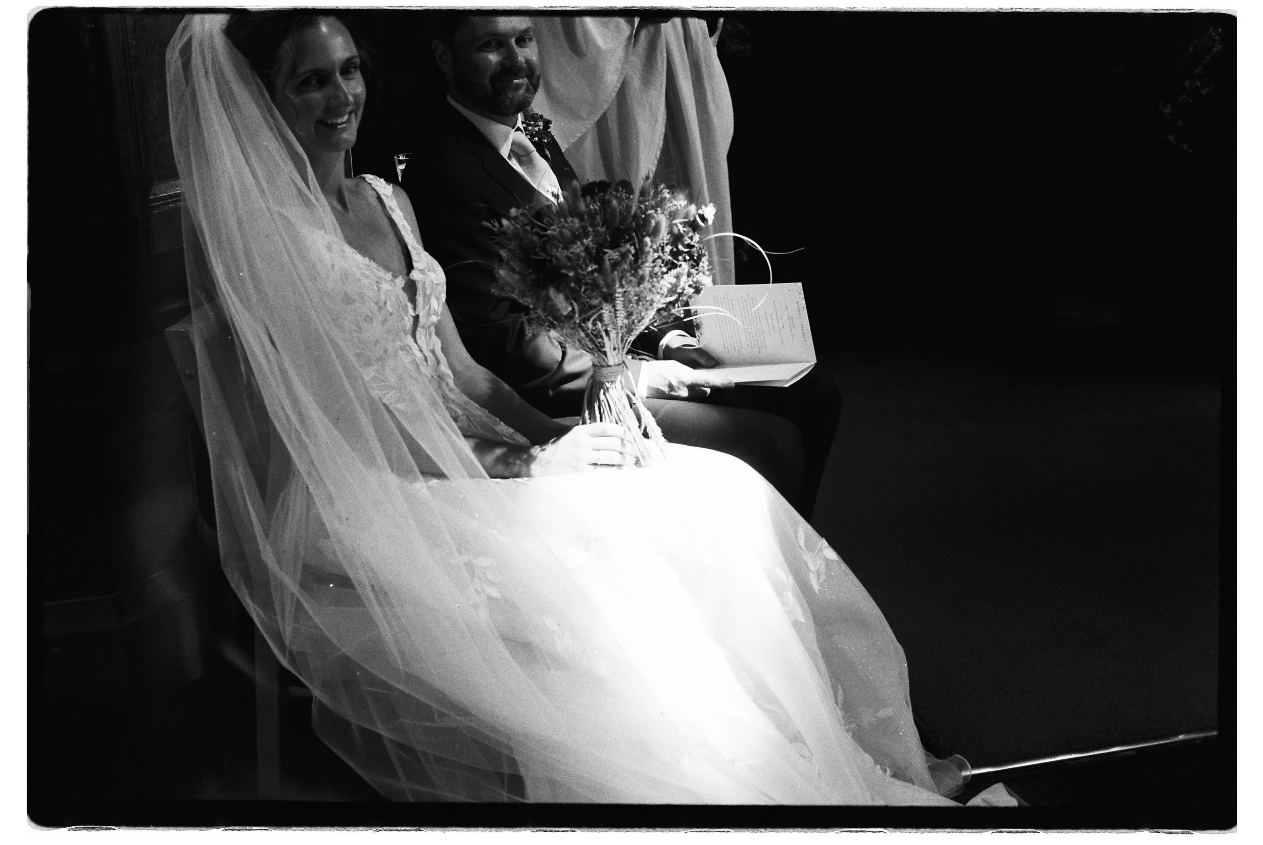 Black and white photo of a bride and groom sitting side by side during a wedding ceremony; the bride is smiling and holding a bouquet of flowers, wearing a wedding dress with a veil, and the groom is smiling, wearing a suit and holding an open book.