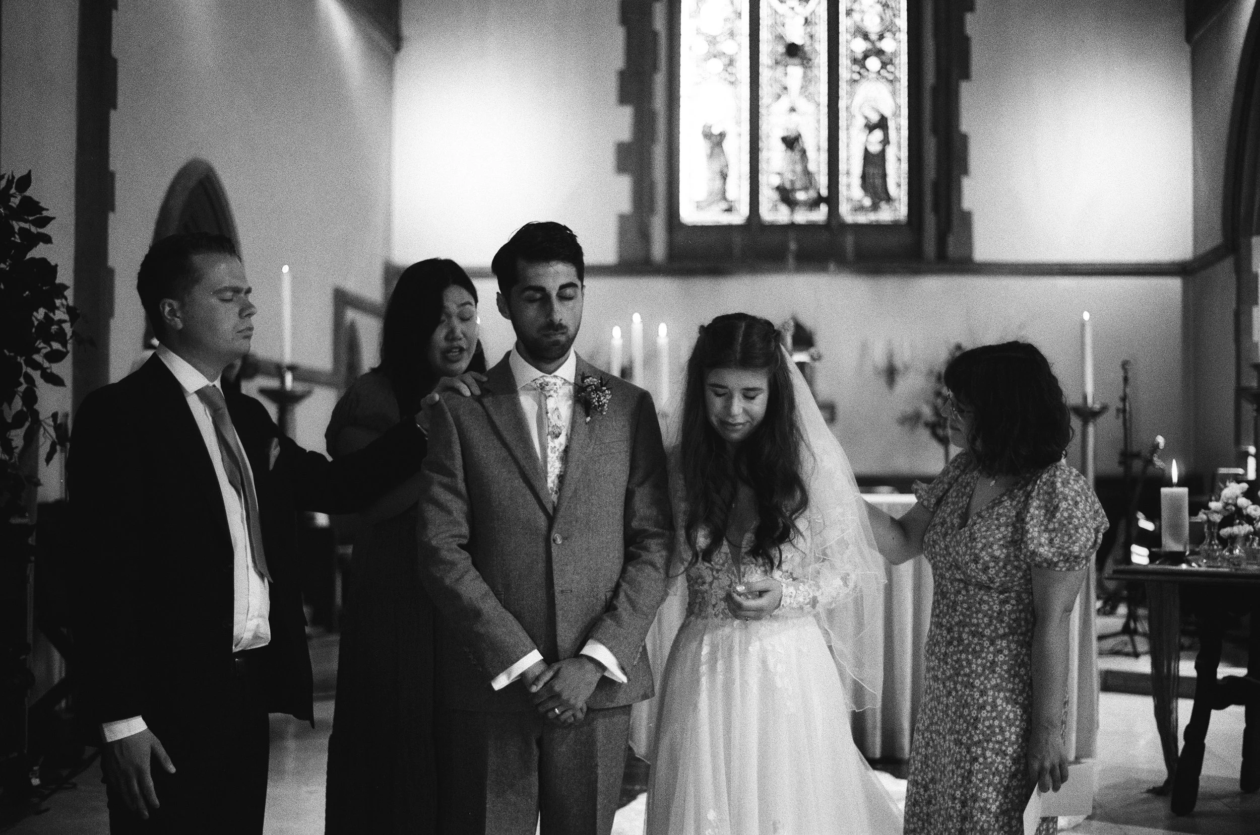 Black and white photo of a wedding ceremony inside a church with five people praying with eyes closed. The bride and groom stand in the center with two women and a young man on either side, all with heads bowed.
