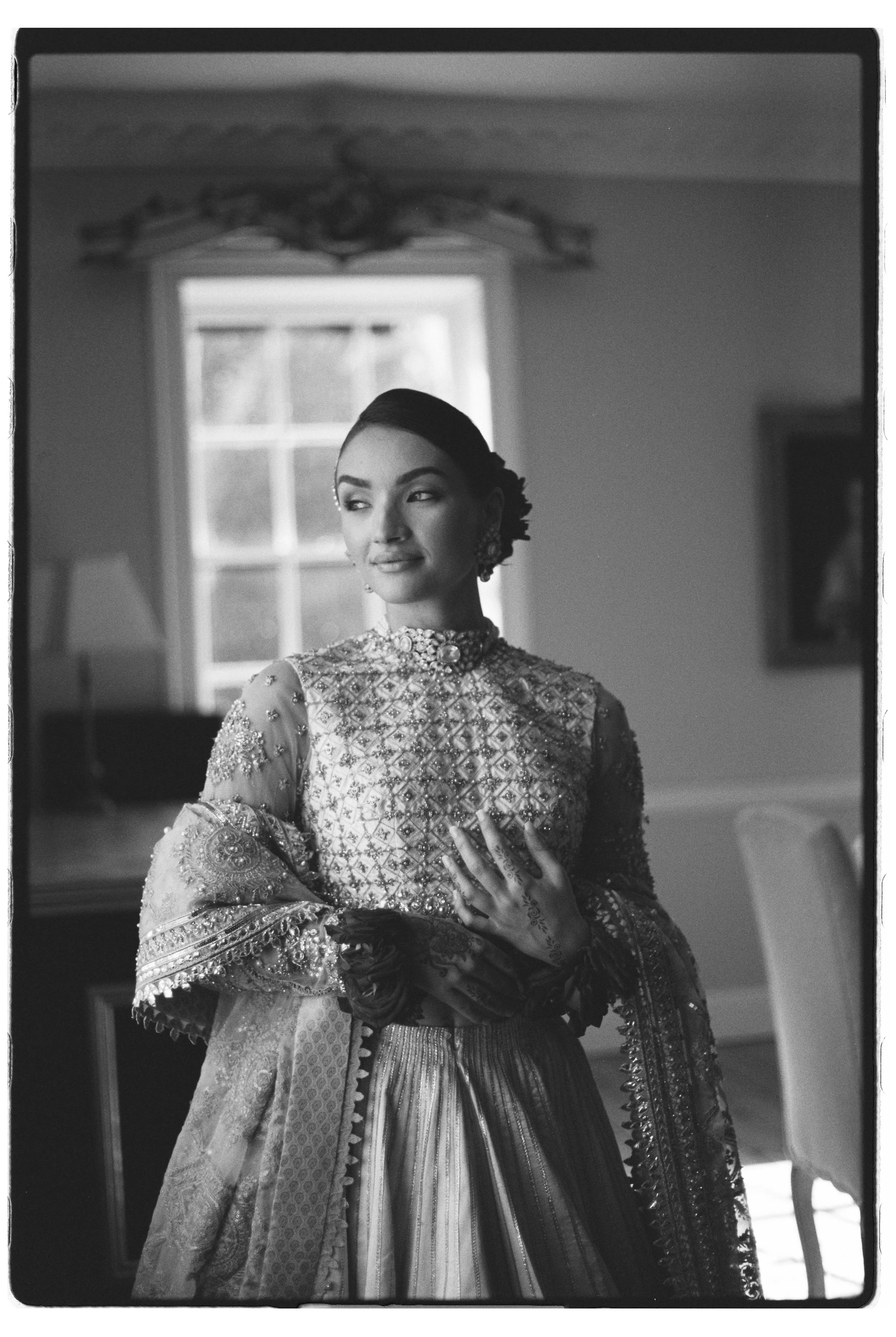 A woman in traditional, ornate attire standing indoors near a window, with her hand on her chest, looking to the side.