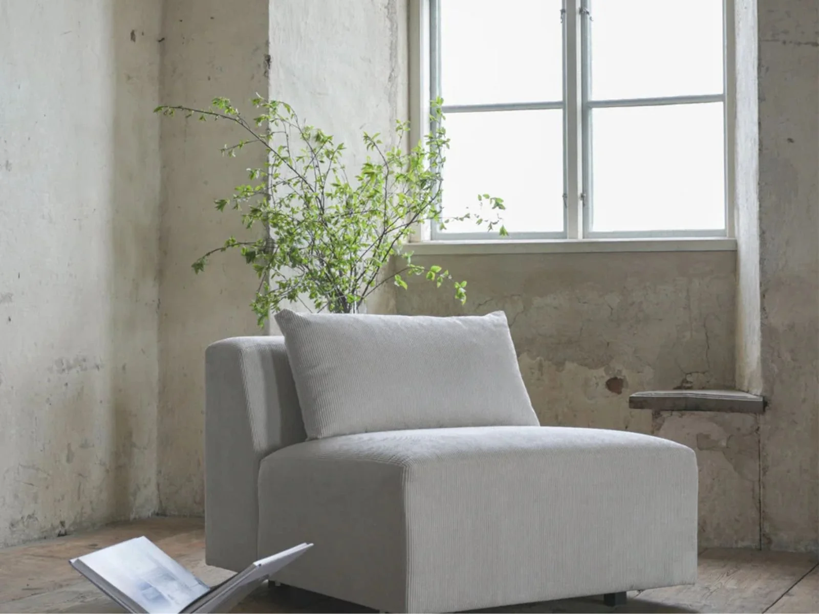 Lyse beige vegger med sprukket og malt overflater, hvit lenestol med pute, grønn plante i bakgrunnen, vindu som slipper inn lys, åpen bok liggende på gulvet.