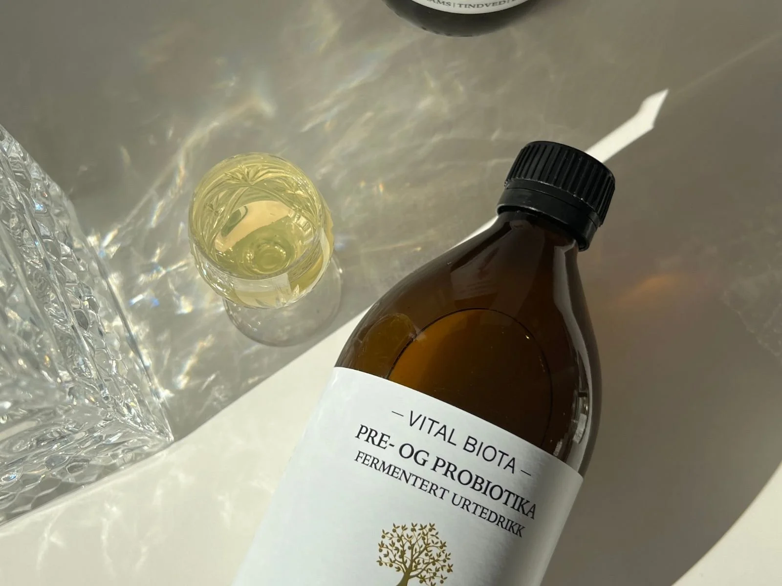 Et brunt glassflaske med hvit etikett og svart lokk, som inneholder pre- og probiotika. Ved siden av en liten, rund, klar glassflaske med gul væske. Begge er plassert på en lys overflate, med en reflekterende overflate bak.