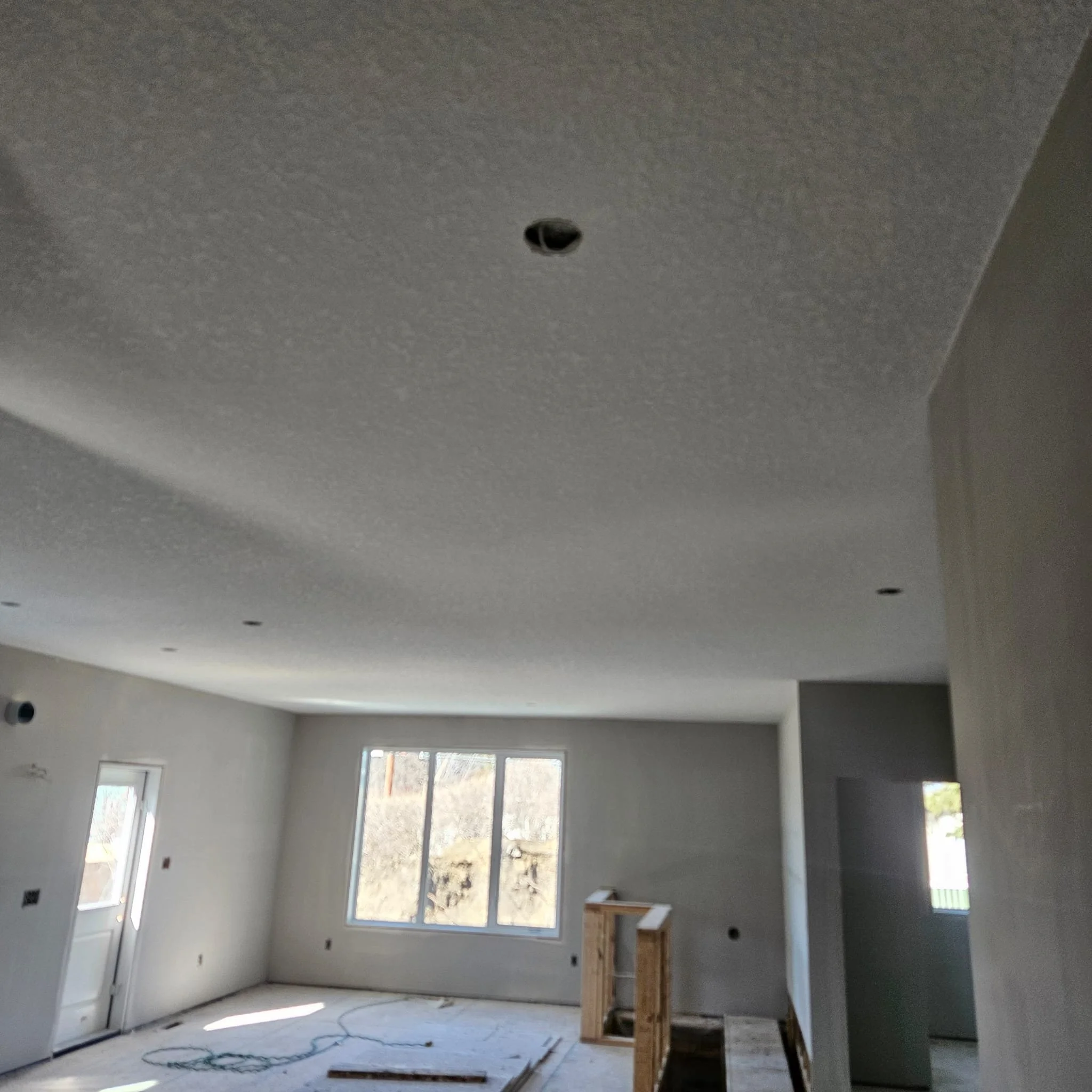 Interior of a room under construction with unpainted drywall, a partially finished ceiling, and an unfinished floor. A rectangular window and partially installed door are visible, along with some construction materials and exposed wires on the floor.
